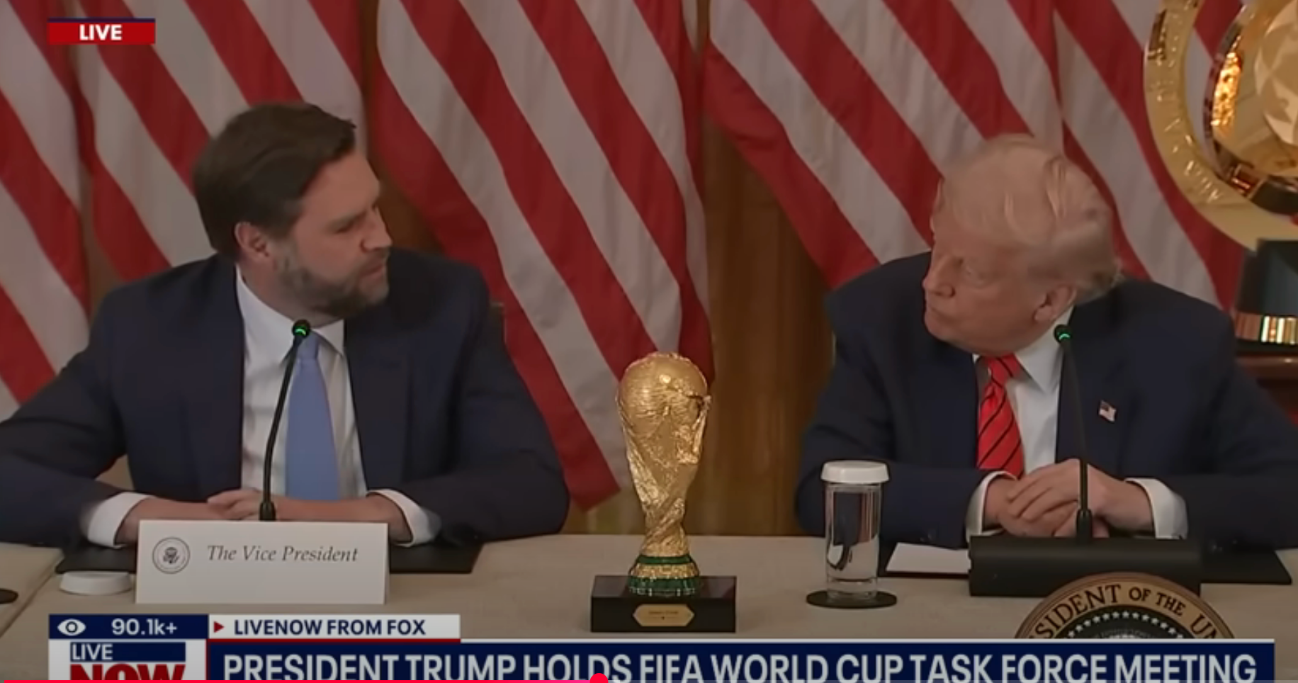 Two men seated at a table with microphones, discussing at a FIFA World Cup Task Force meeting, with a World Cup trophy in front of them