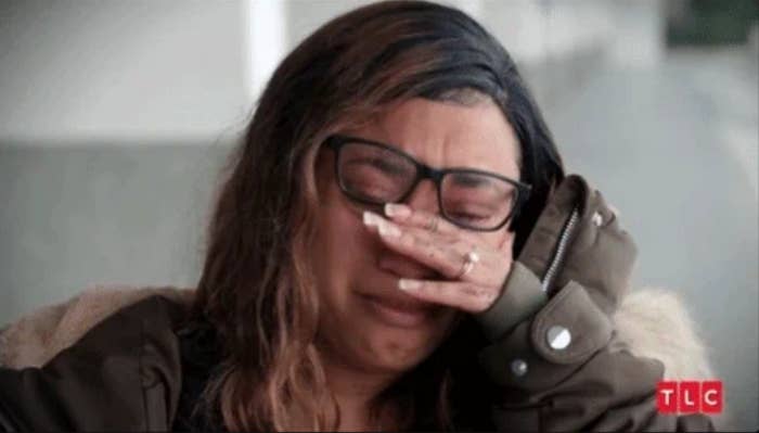Person with glasses and long hair crying, covering their face with one hand while seated indoors, conveying emotions related to work challenges