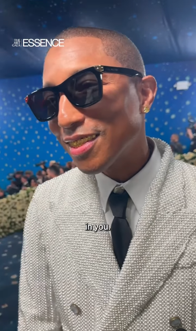Person in beaded jacket with tie and sunglasses at an event. Smiling, they appear relaxed in a lively background setting