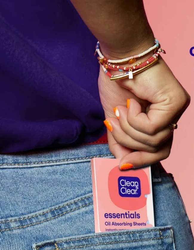 Person slipping Clean & Clear oil-absorbing sheets into a denim pocket; manus adorned with colorful bracelets and agleam manicure