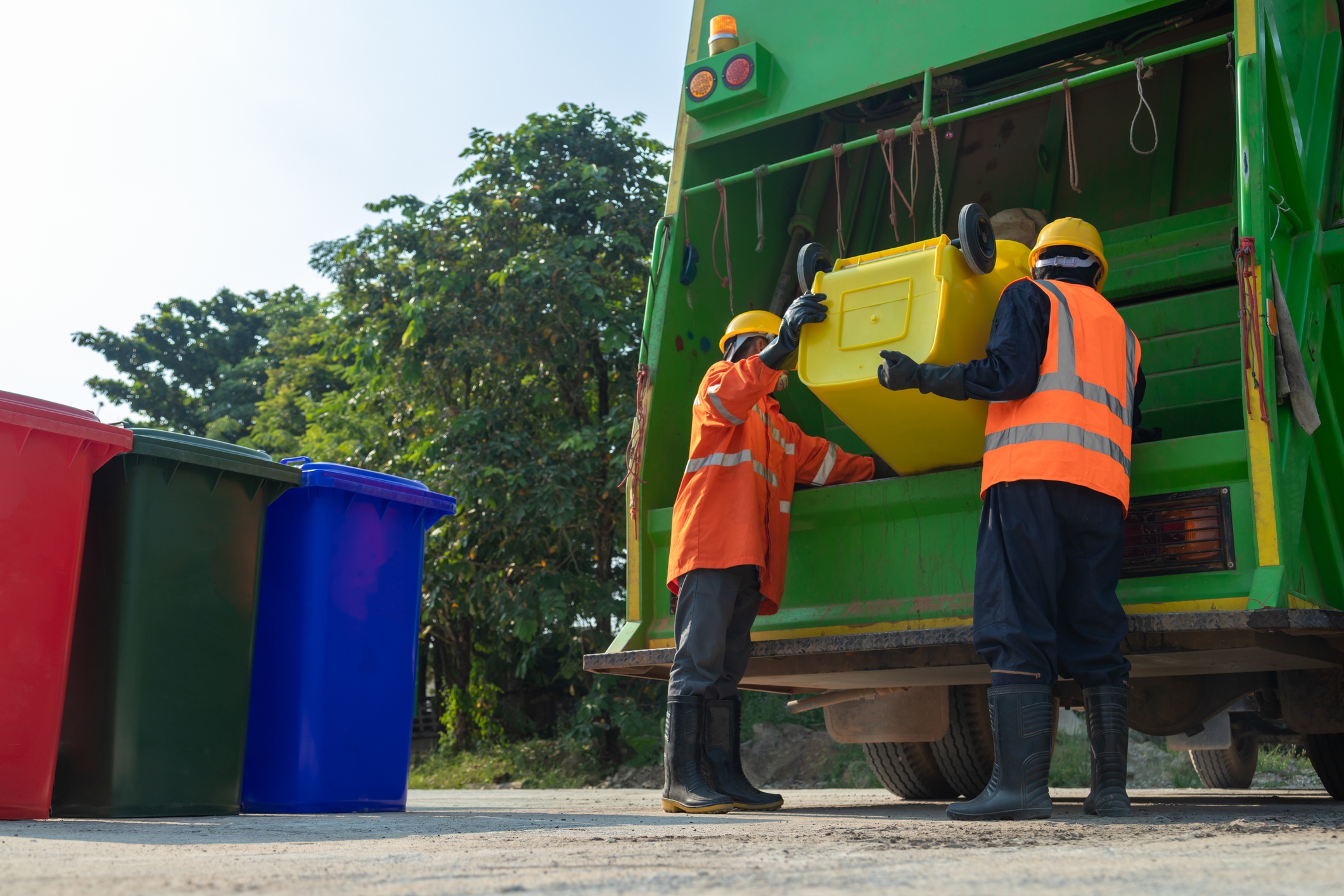 Two sanitation workers in uniform load a yellow bin into a green garbage truck, with red, blue, and green bins nearby