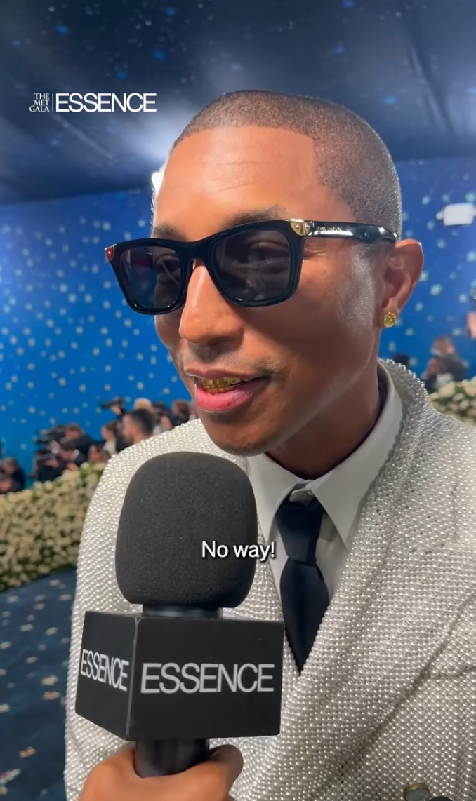 Person in sparkly suit and sunglasses being interviewed at a formal event, holding an ESSENCE microphone, under a blue starry backdrop