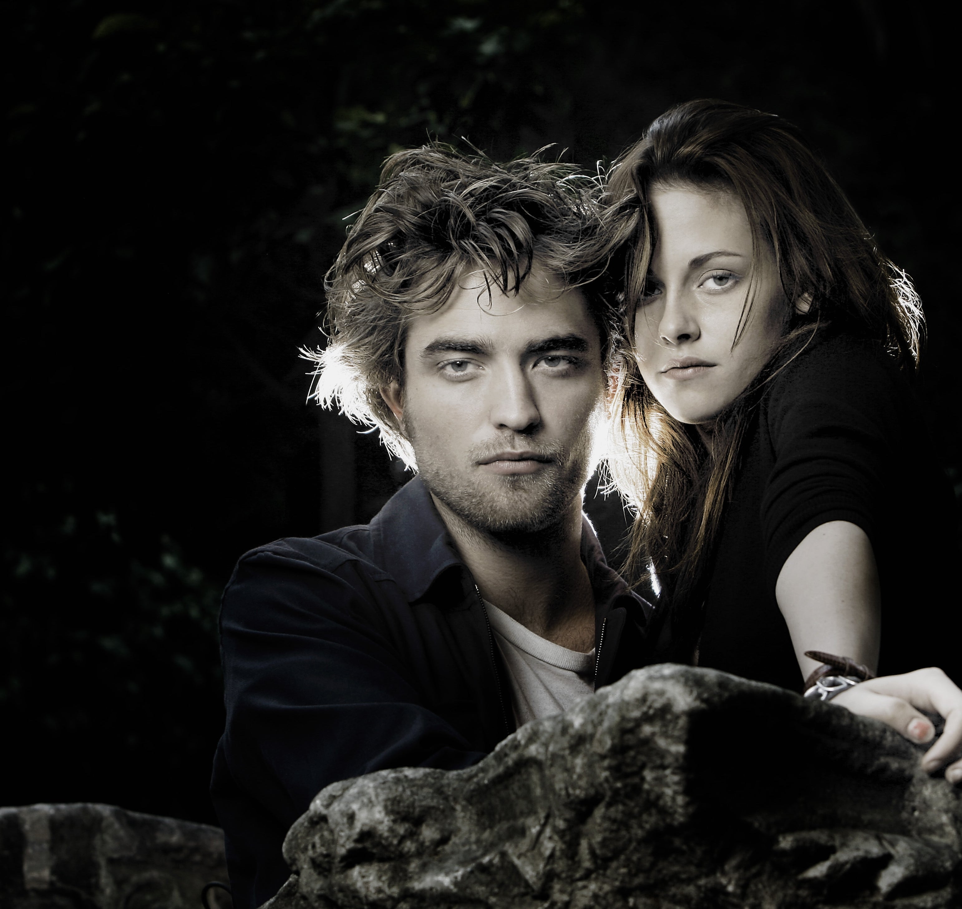 Two people stand close together, gazing seriously at the camera, in an outdoor setting