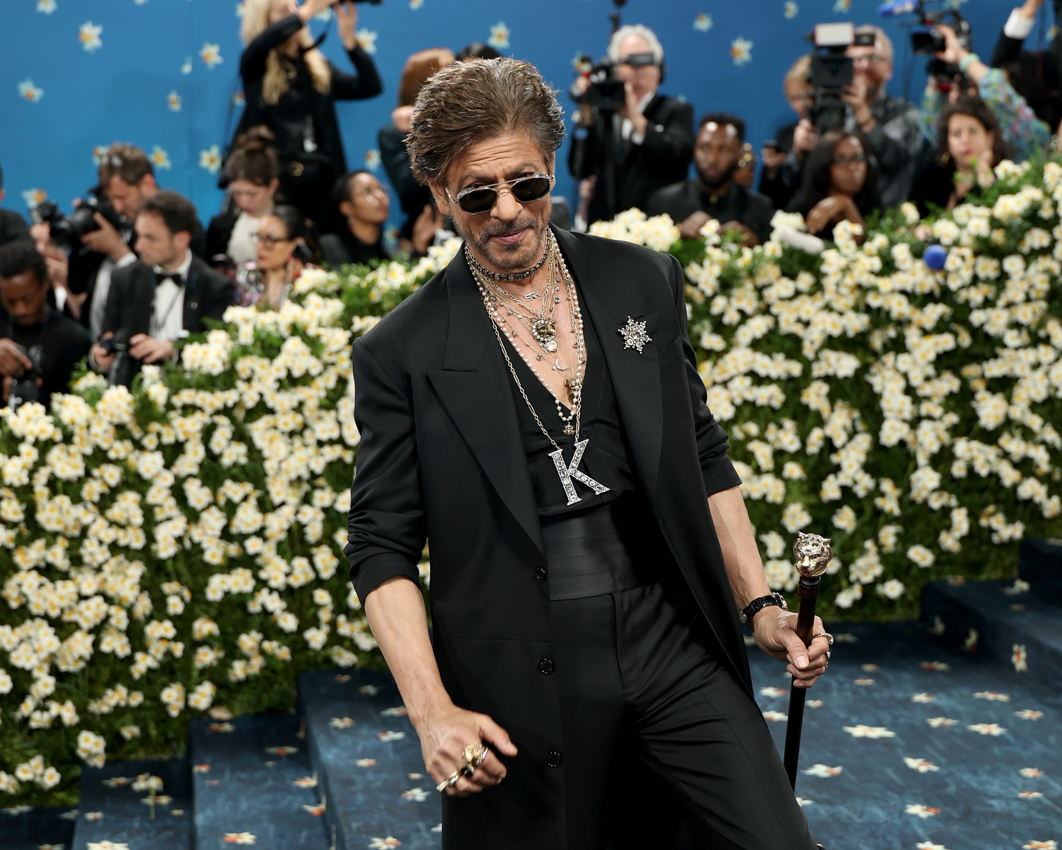 A celebrity on a red carpet wears a sleek black suit, layered necklaces, and sunglasses, holding a decorative cane, surrounded by photographers