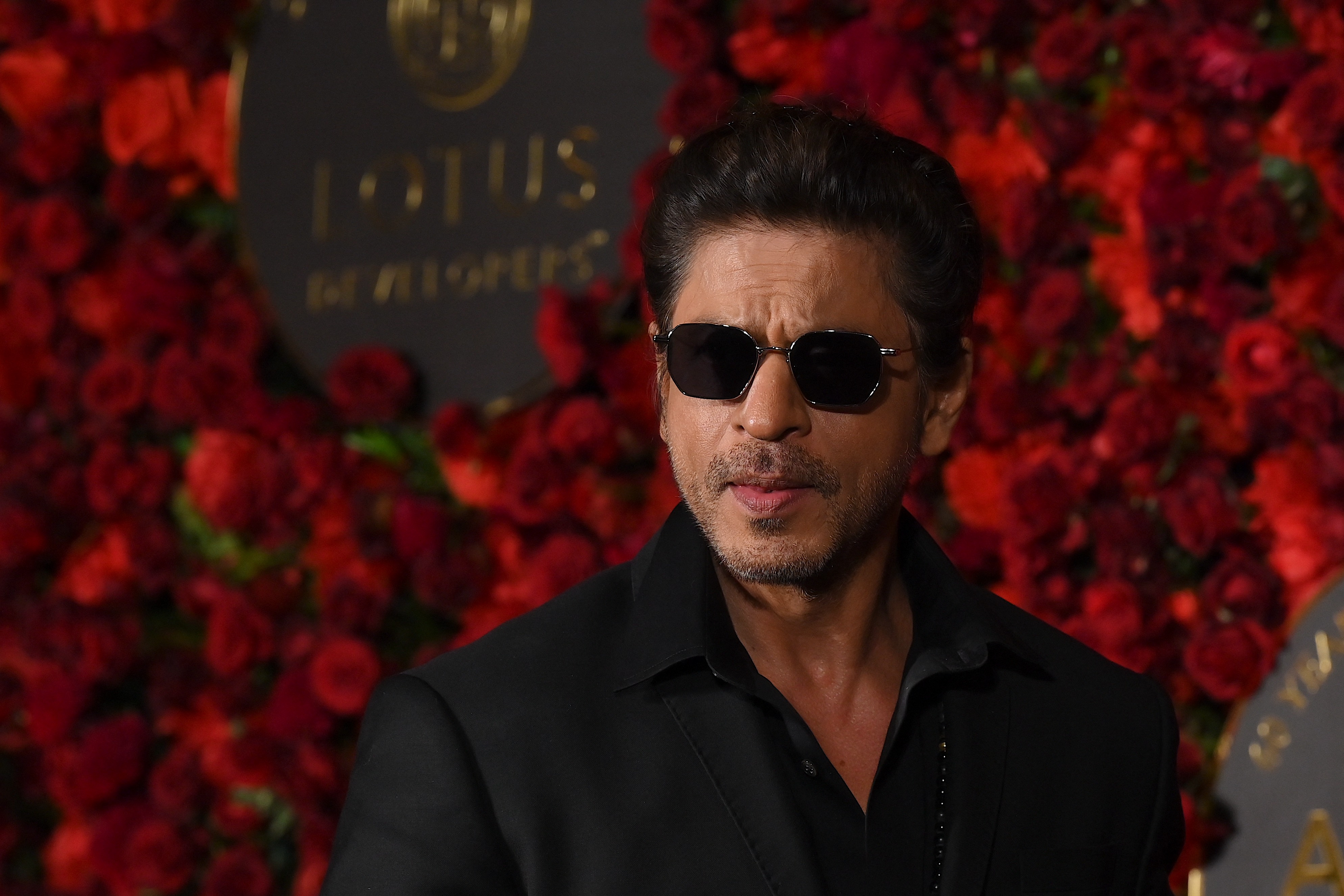 A person in sunglasses on a red carpet, wearing a dark, formal suit with a confident expression, surrounded by a backdrop of red roses