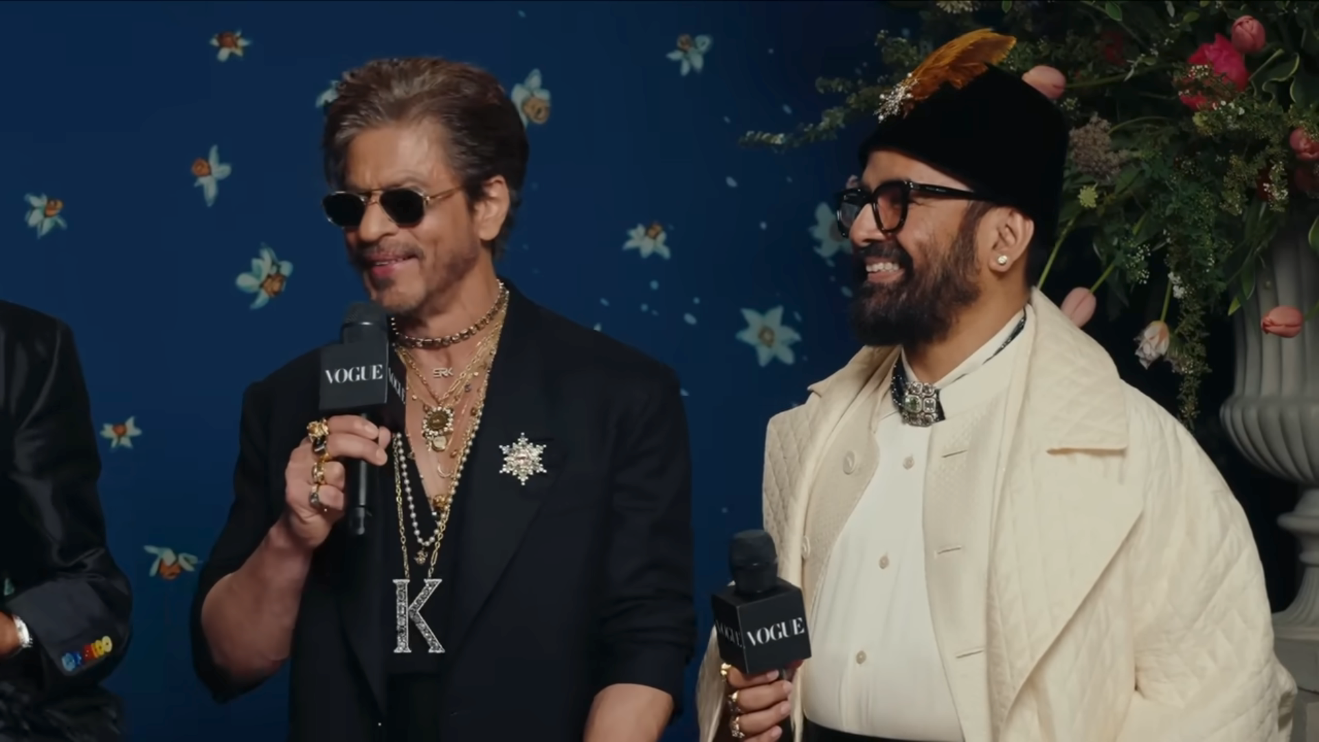Two men on a red carpet, both holding microphones. One wears a black suit with layered necklaces and sunglasses; the other is in a white coat and hat
