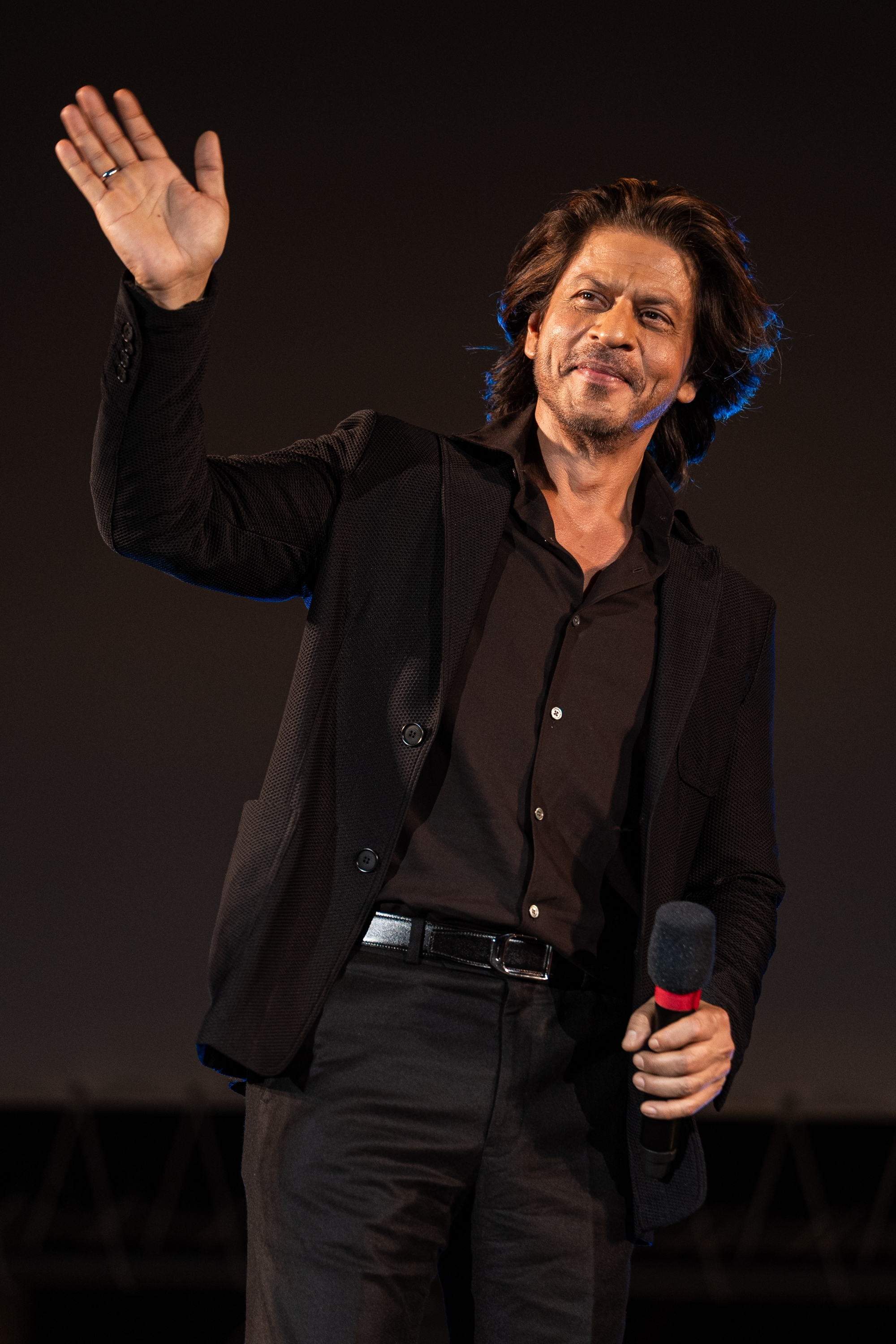 Person in a dark suit waving and holding a microphone, in a public appearance setting