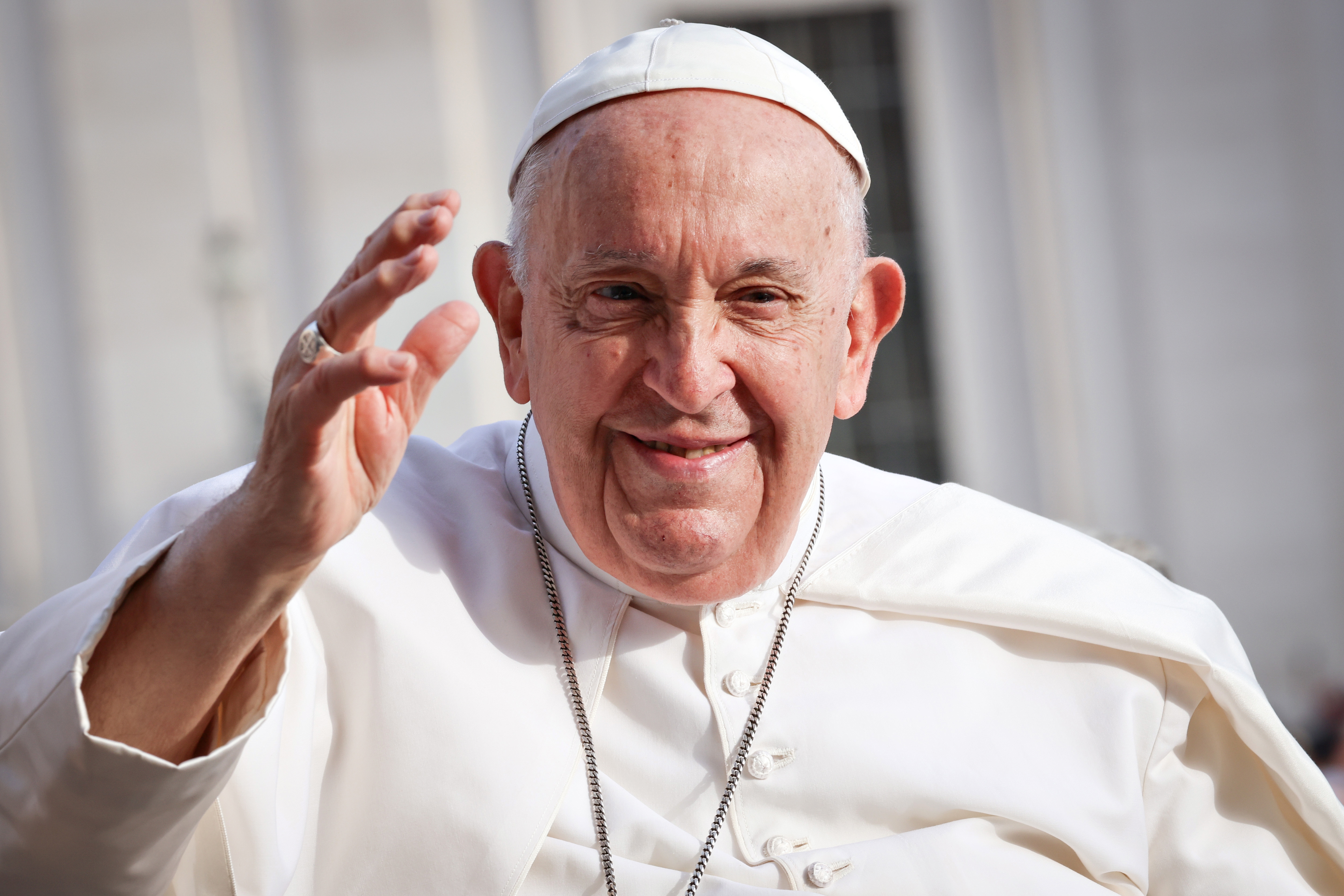 A religious leader with a white cassock and cap waves outdoors, smiling warmly