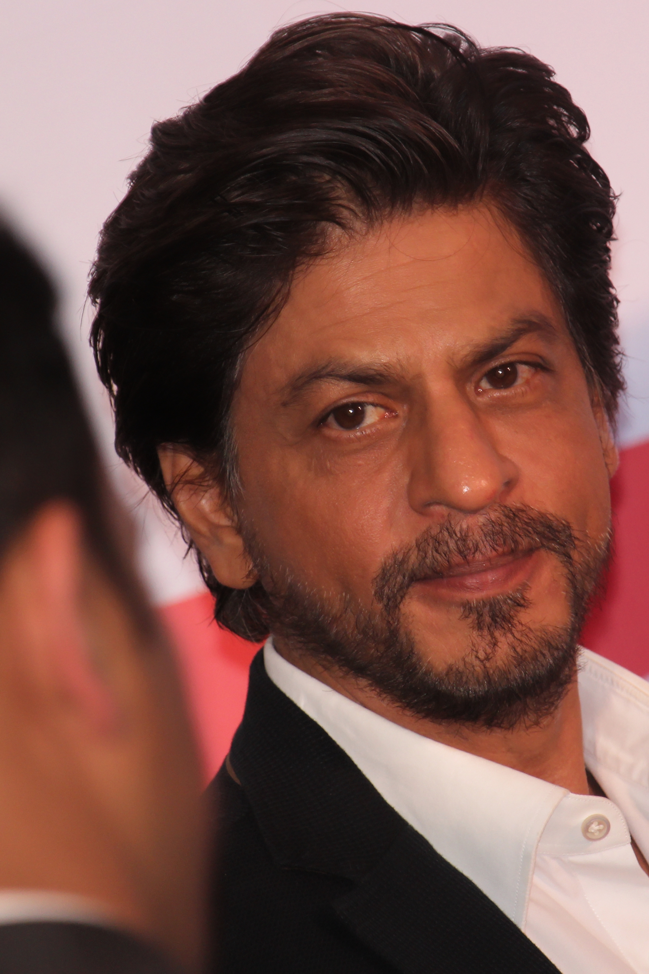 A person with wavy hair and a beard, wearing a formal suit, attends an event