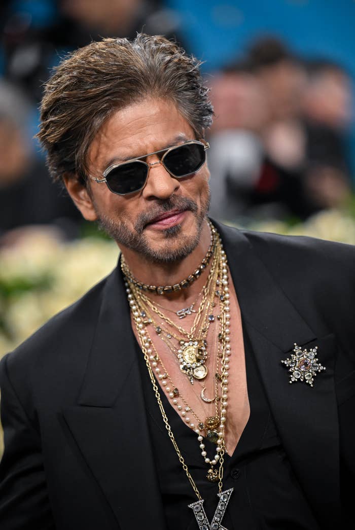 Man wearing layered gold necklaces, sunglasses, and a black suit with an embellished brooch, smiling at a celebrity event