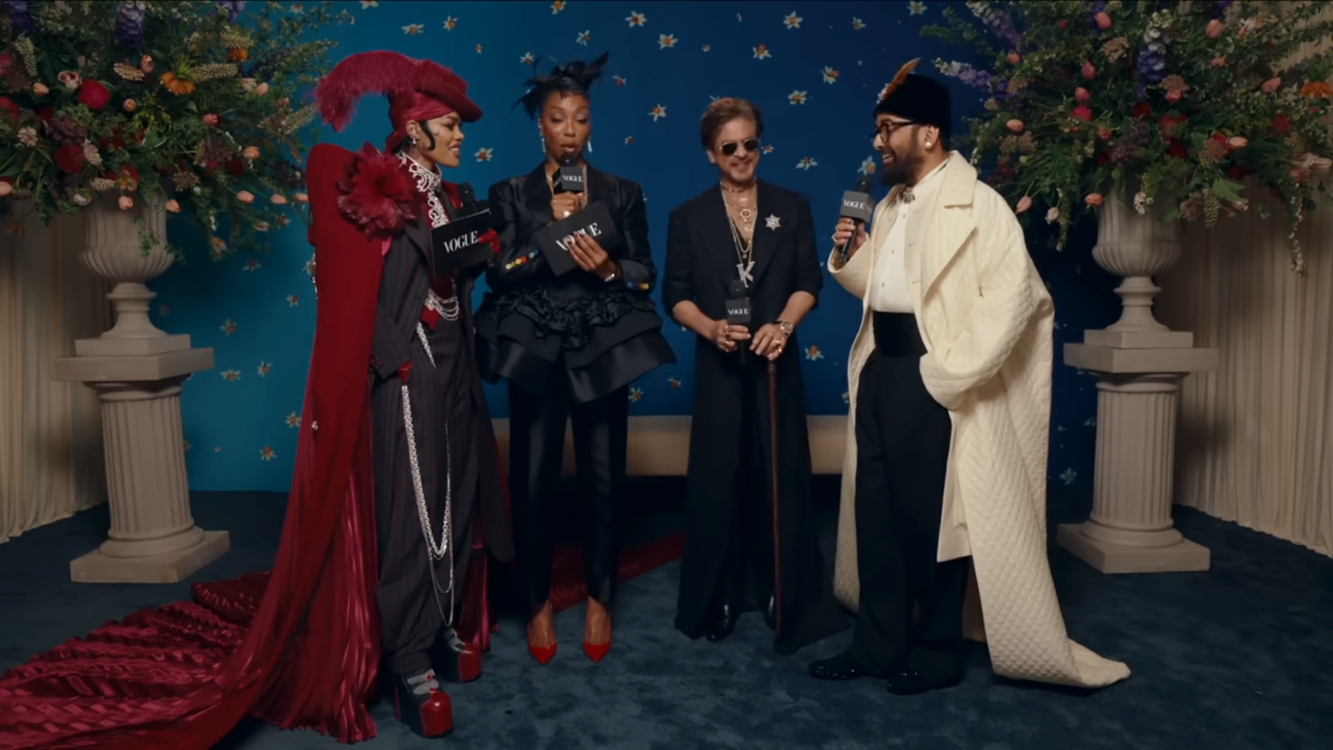 Four individuals in extravagant, formal outfits engage in an interview standing amidst floral decor