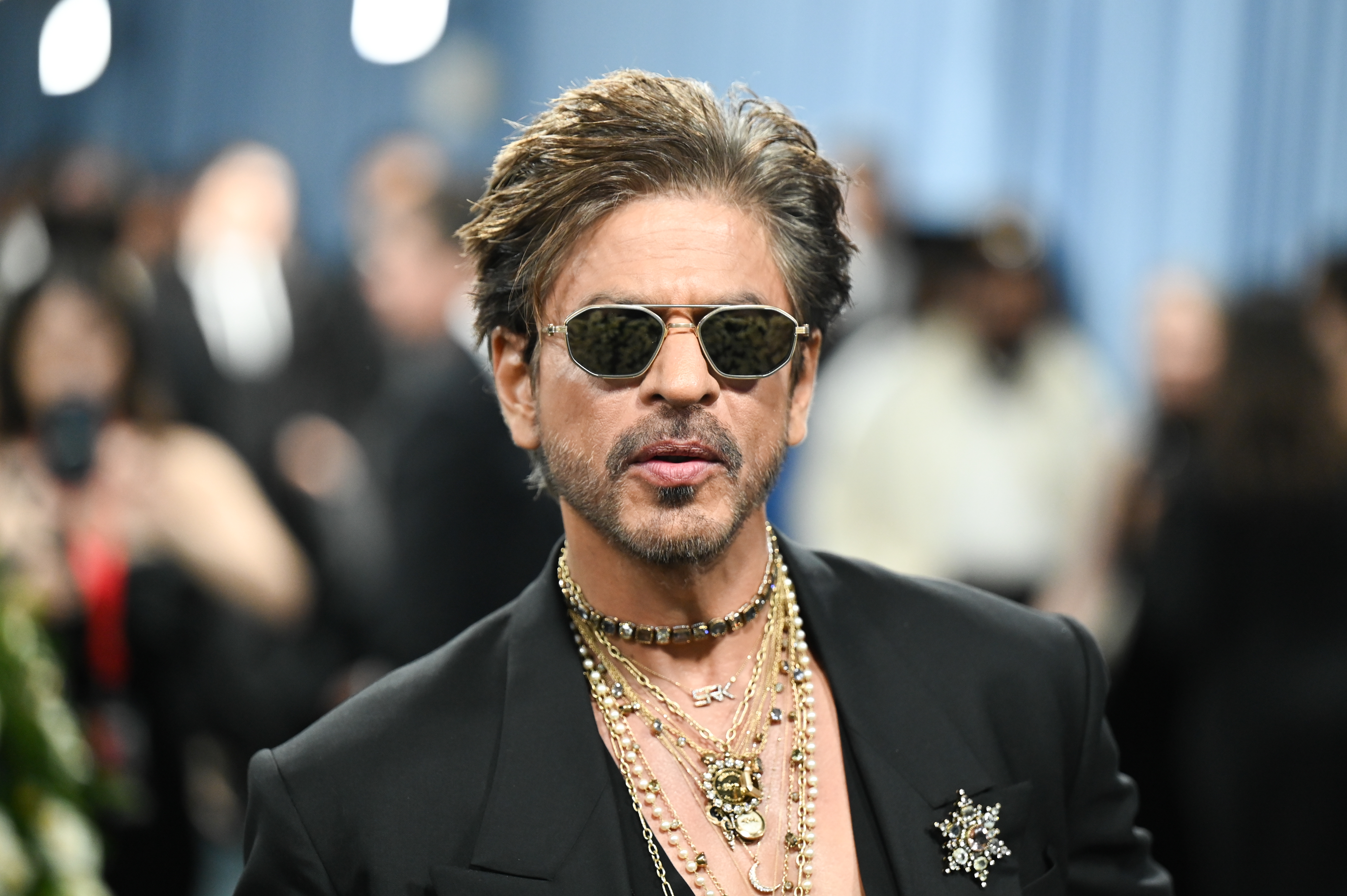 A man in a stylish black suit adorned with layered gold necklaces and sunglasses attends a formal event