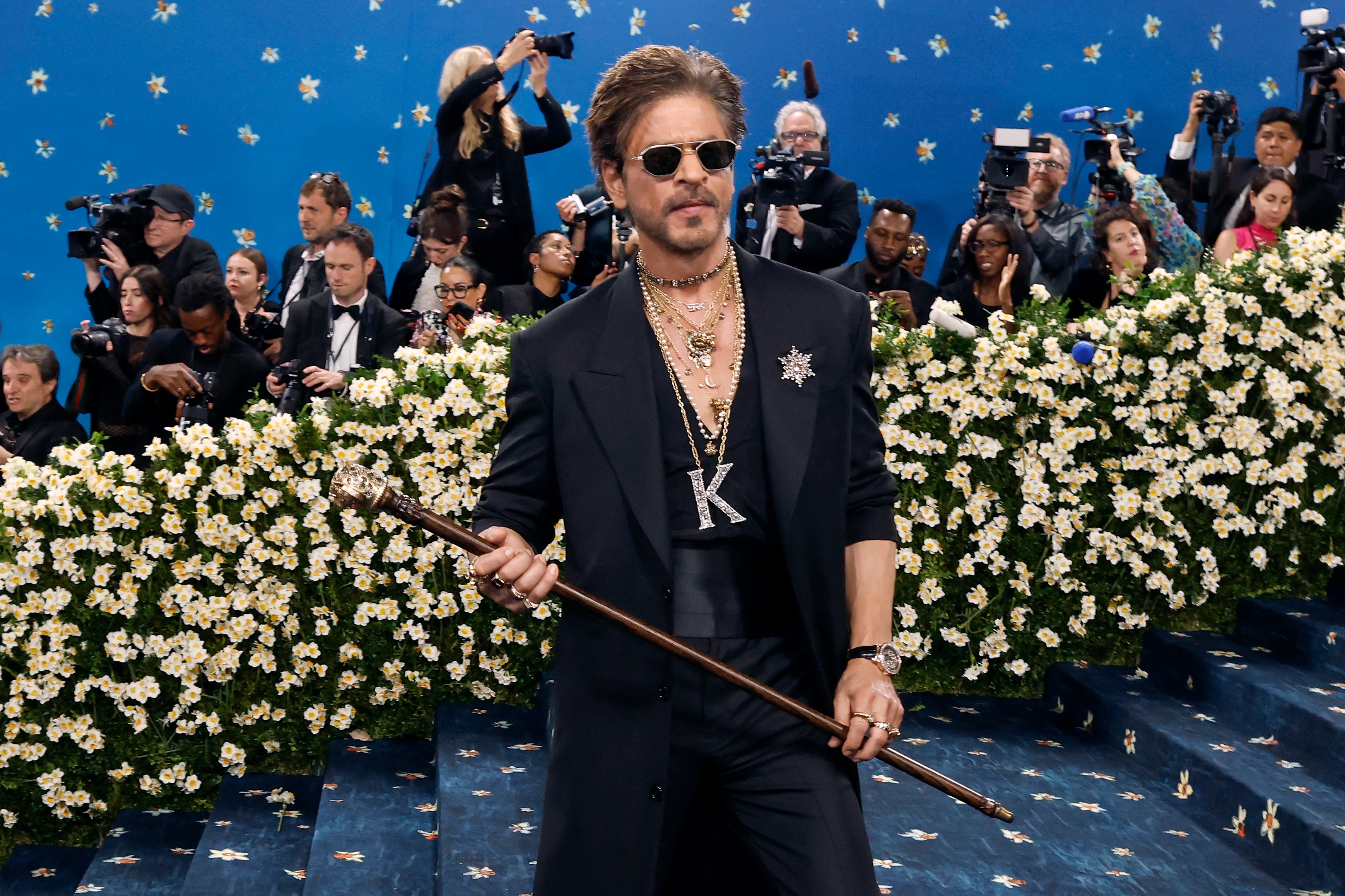 Person wearing a dark suit and sunglasses, holding a cane, surrounded by photographers and flowers at a celebrity event