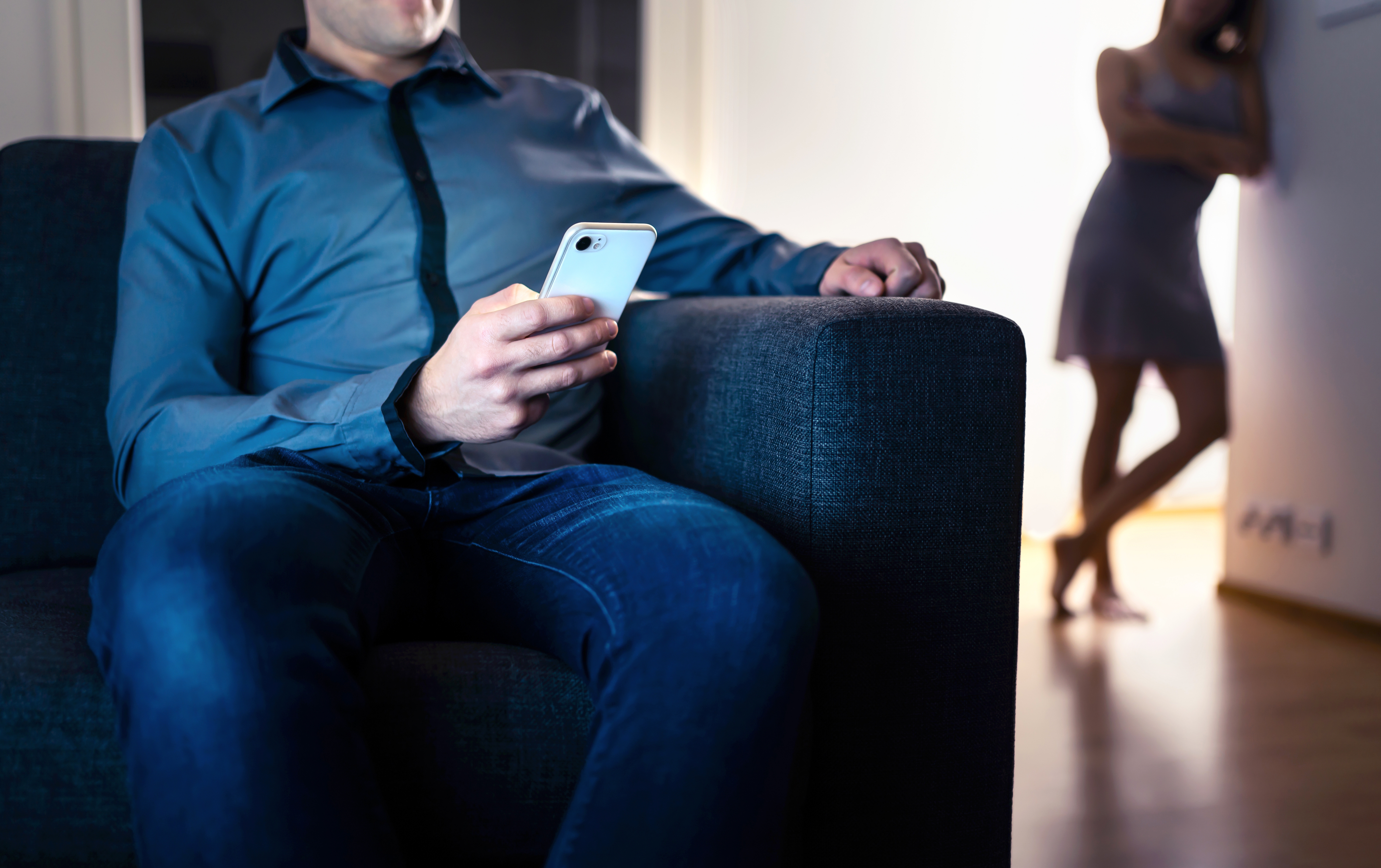 Man on phone seated, while a woman stands in the background, suggesting a potential relationship tension