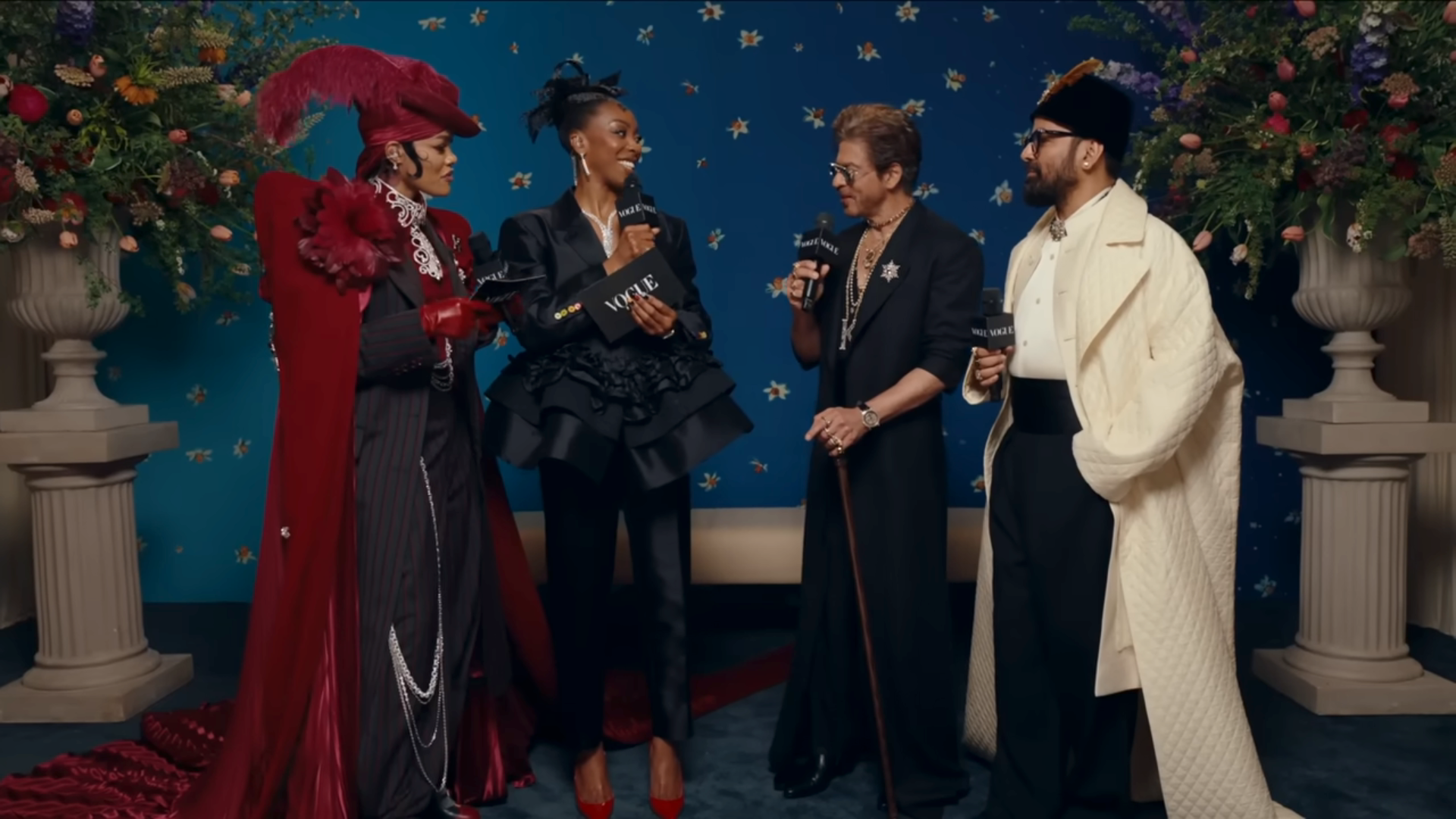 Four people in elegant, high-fashion outfits chat on a red carpet. Bold accessories and dramatic styling highlight their unique fashion choices
