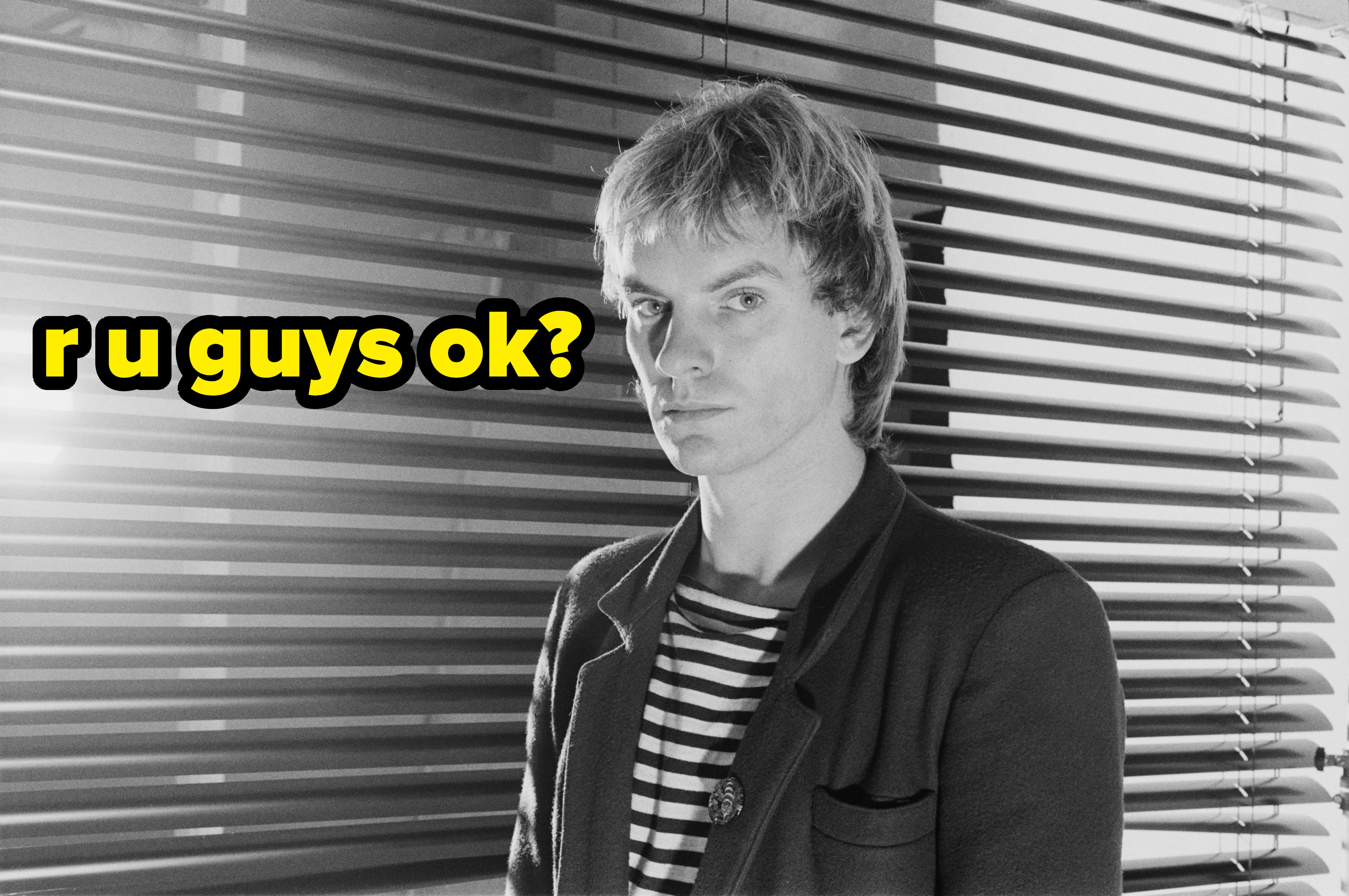 Person in a striped shirt and jacket stands indoors by window blinds, gazing directly at the camera