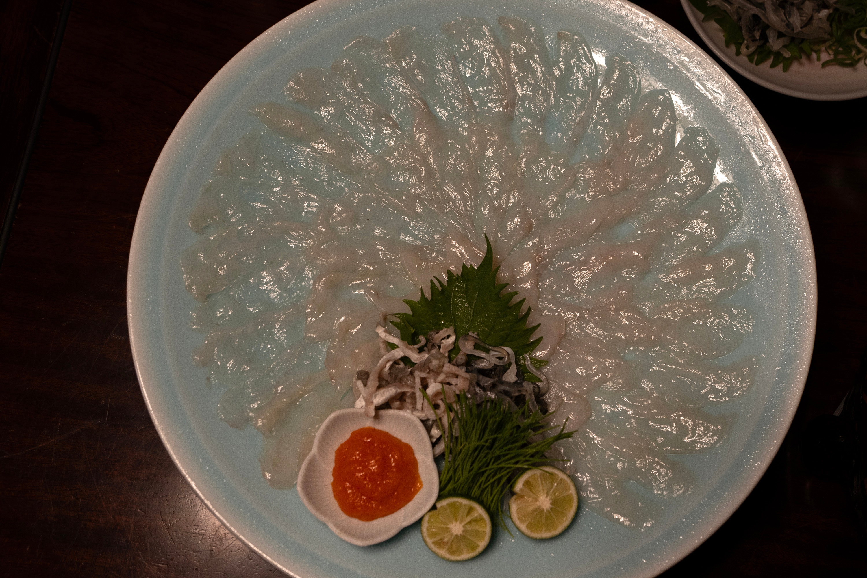Thinly sliced sashimi arranged in a floral pattern on a plate, garnished with greens, lime slices, and a small dish of orange sauce