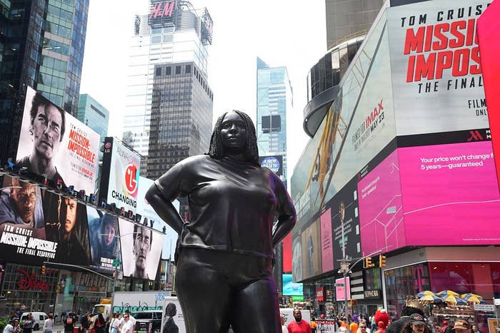 A large, dark sculpture of a woman stands prominently in Times Square, surrounded by numerous digital billboards