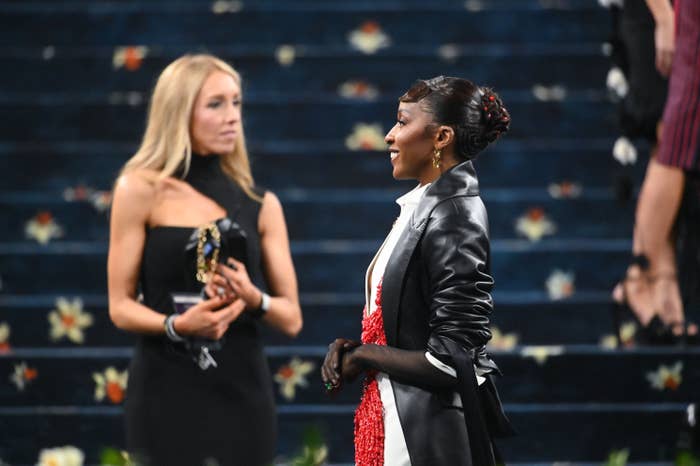 Two individuals on a formal occasion; one in a black dress, the other in a stylish outfit with a long coat and gloves, standing on patterned steps