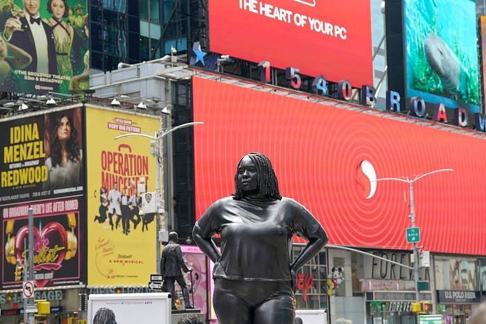 Statue of a woman in Times Square with theatre ads and a large tech billboard in the background