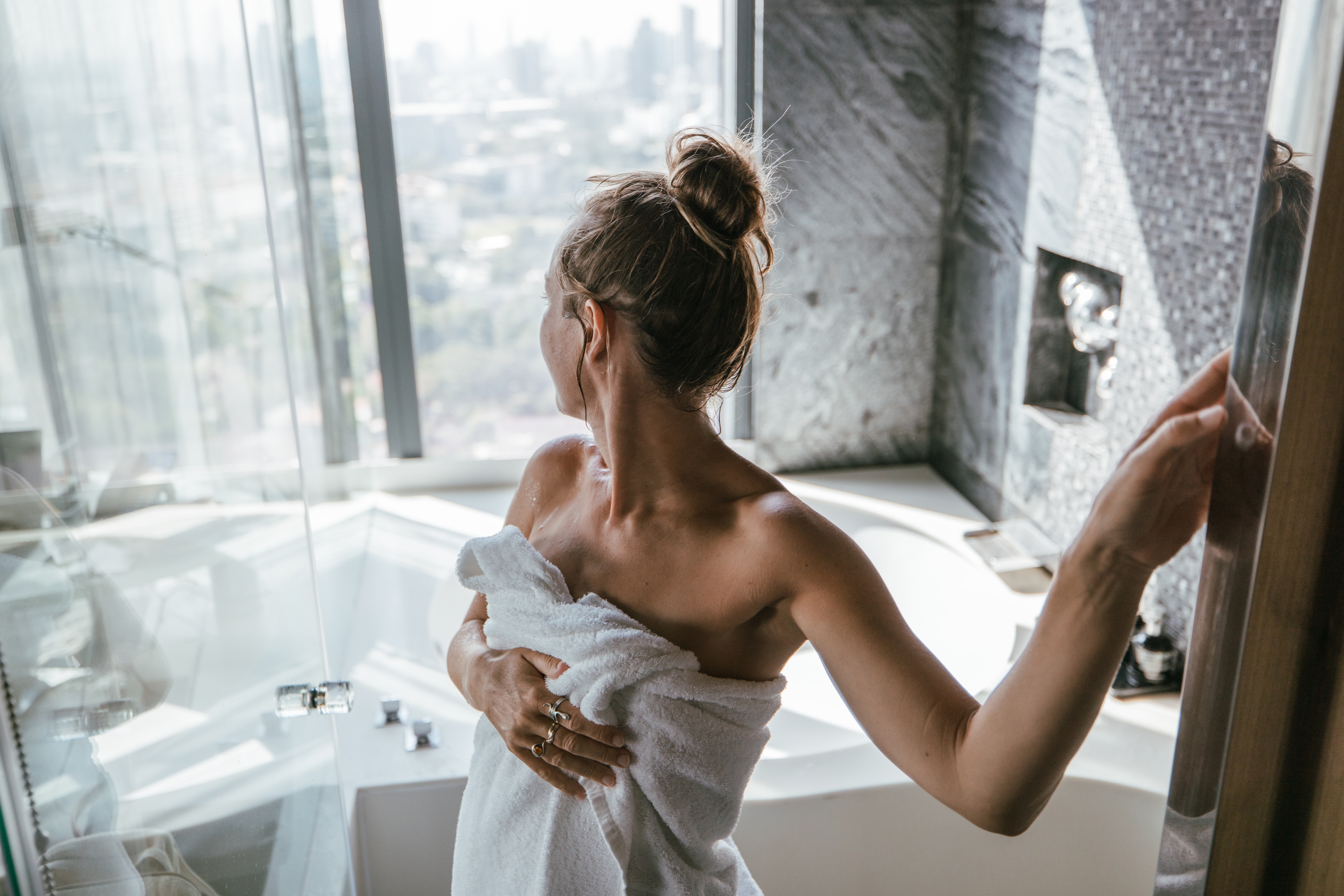 Person wrapped in a towel looks out a large window in a modern bathroom, revealing a cityscape view