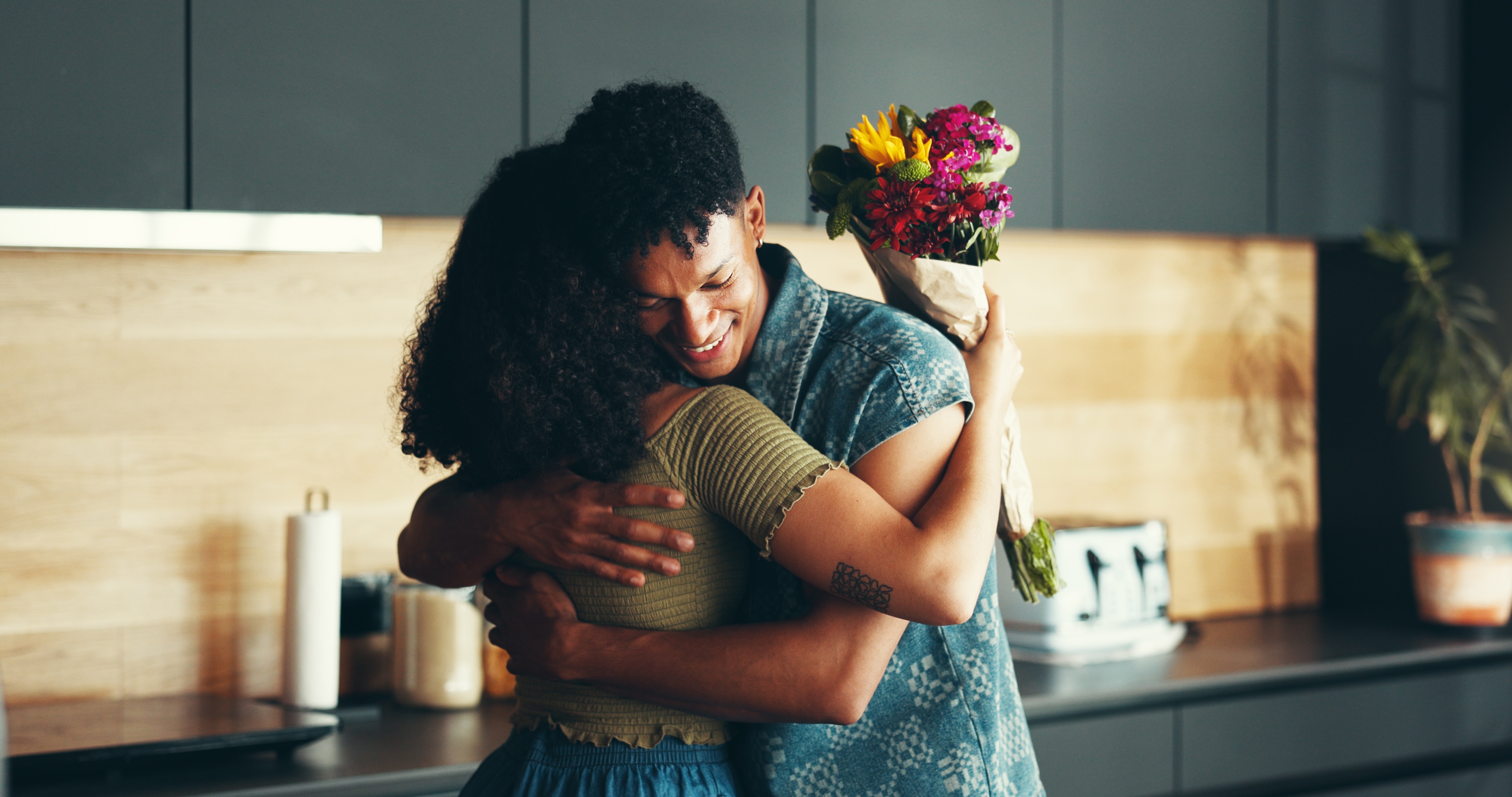 A couple embraces lovingly in a kitchen. One person holds a bouquet of flowers, signifying affection and togetherness