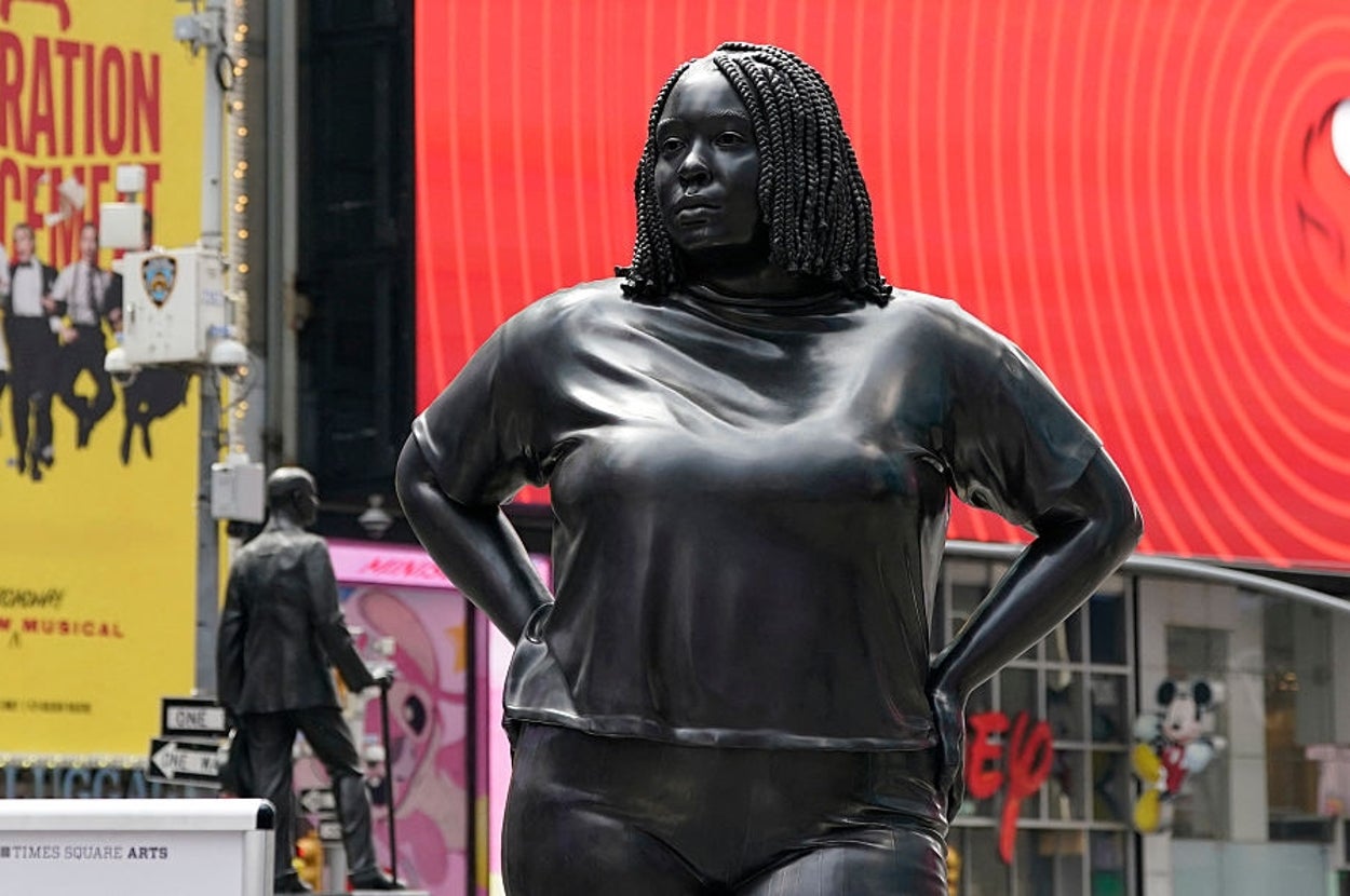 A sculpture of a woman with beaded hair stands confidently in Times Square, surrounded by billboards and city signage