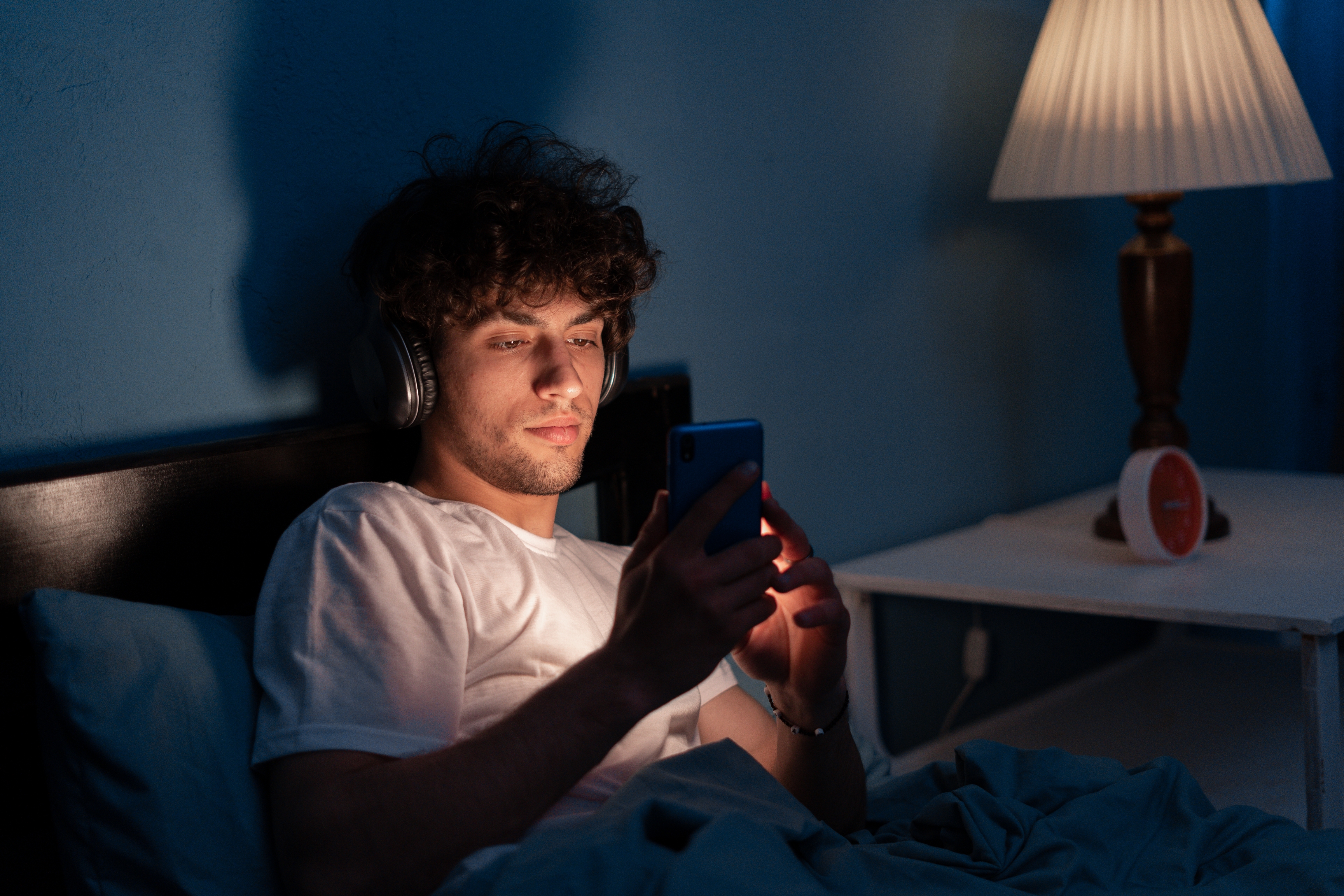 Man in bed wearing headphones, looking at a smartphone with a thoughtful expression in a dimly lit room