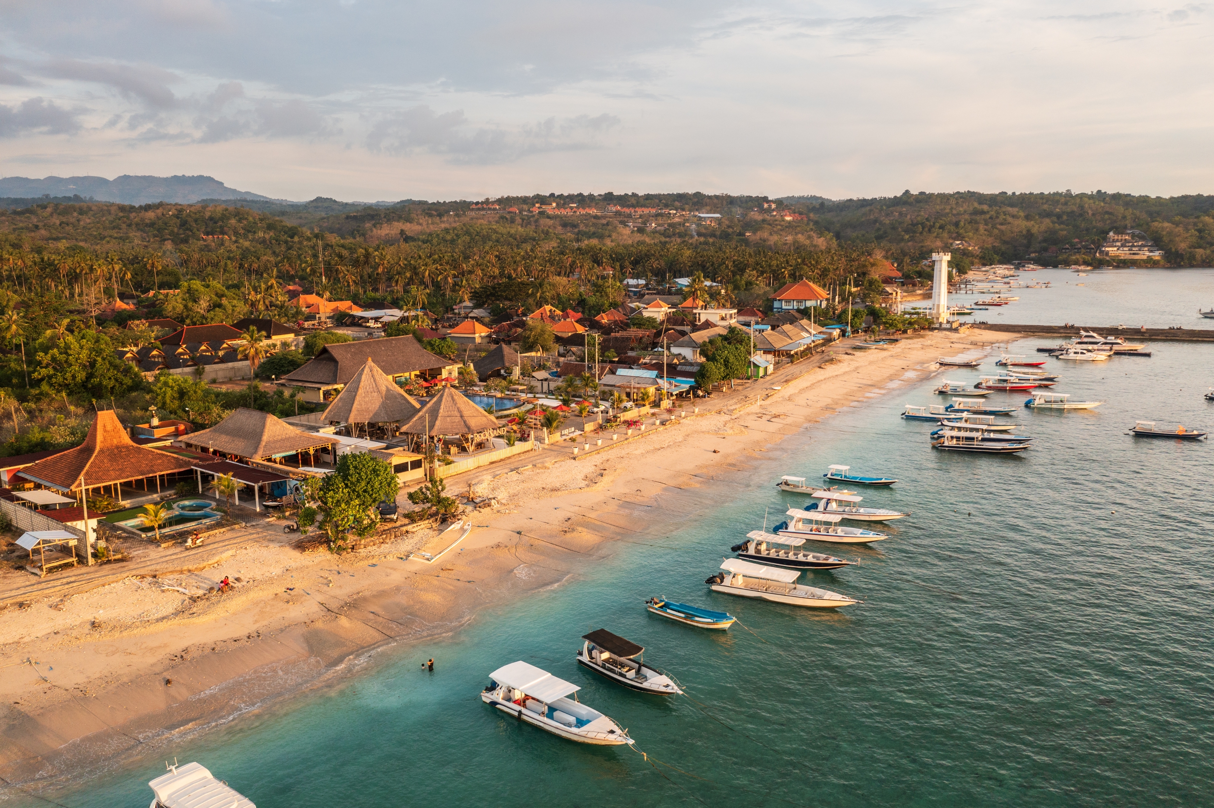 Aerial view of a tropical beach with boats lined up on the shoreline, adjacent to a village with traditional-style buildings and a distant lighthouse