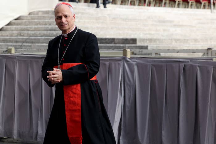 A clergyman wearing formal religious attire with a red sash stands outdoors on stone steps