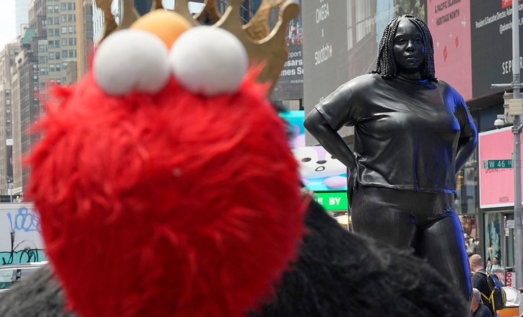 Close-up of a person in an Elmo costume on a busy street, with a statue of a woman in casual clothes in the background