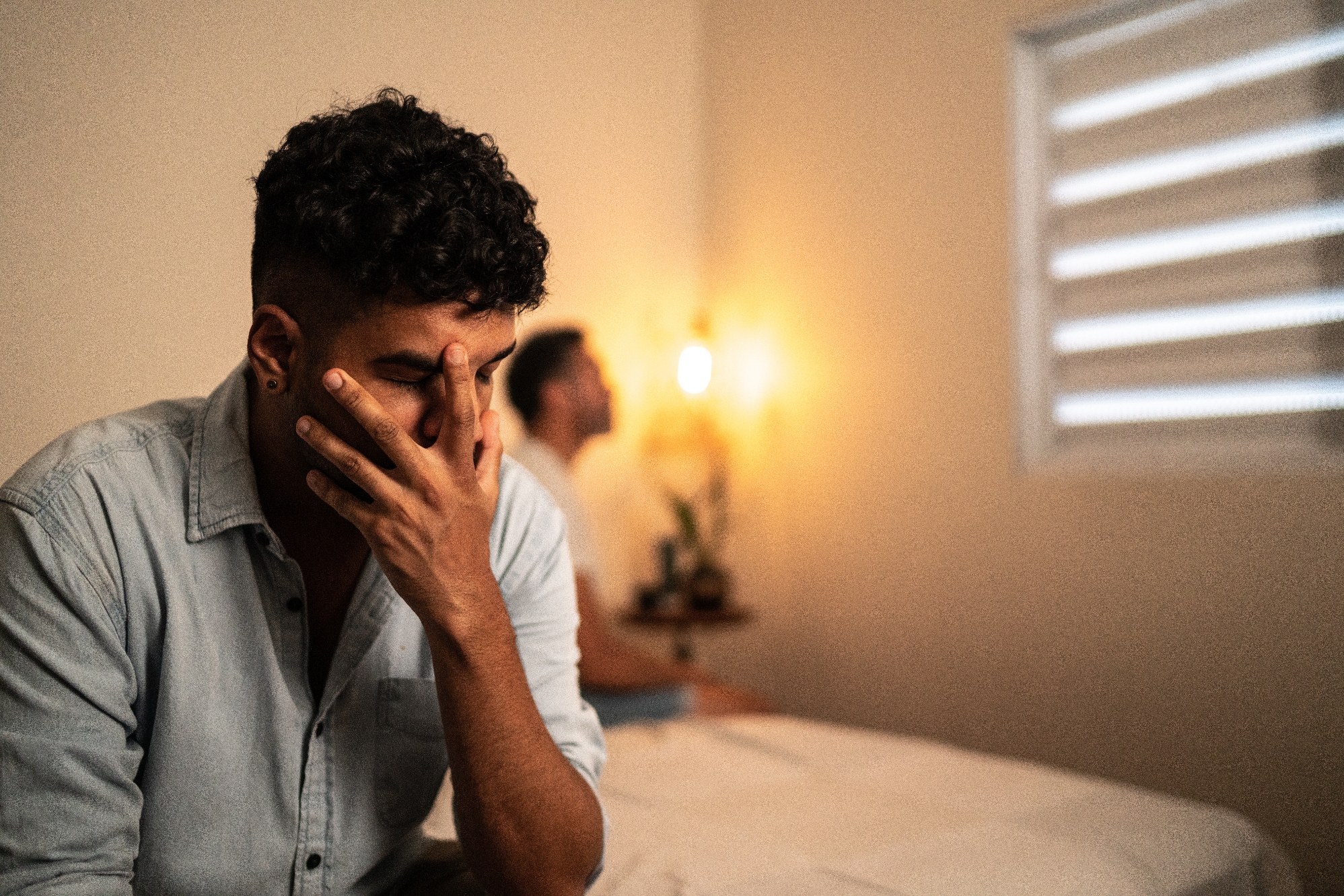 Man sitting on bed with hand on face, appearing upset. Another person is blurred in the background, sitting with their back turned