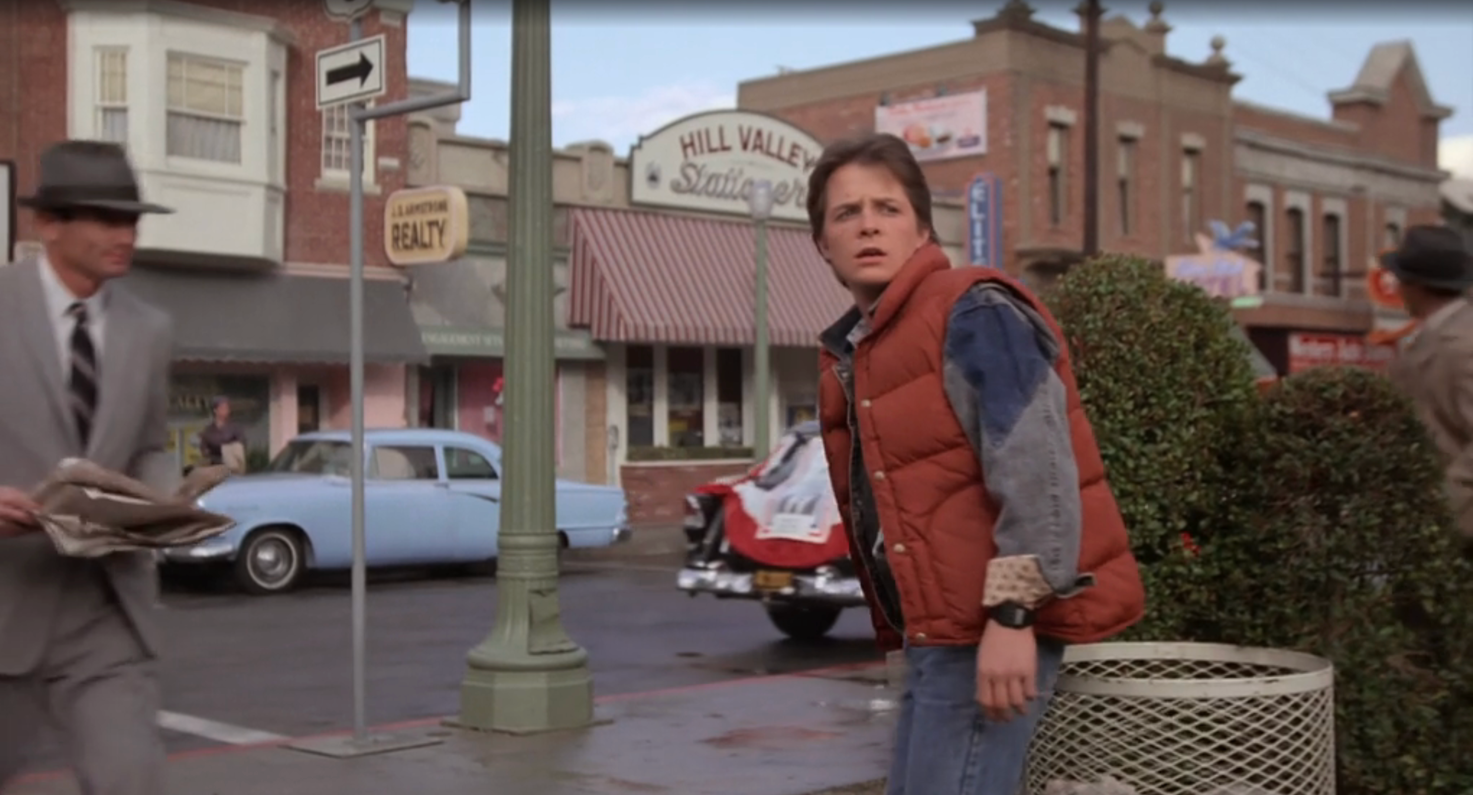 Michael J Fox in a denim jacket and vest looks startled on a 1950s-style street, with vintage cars and buildings in the background