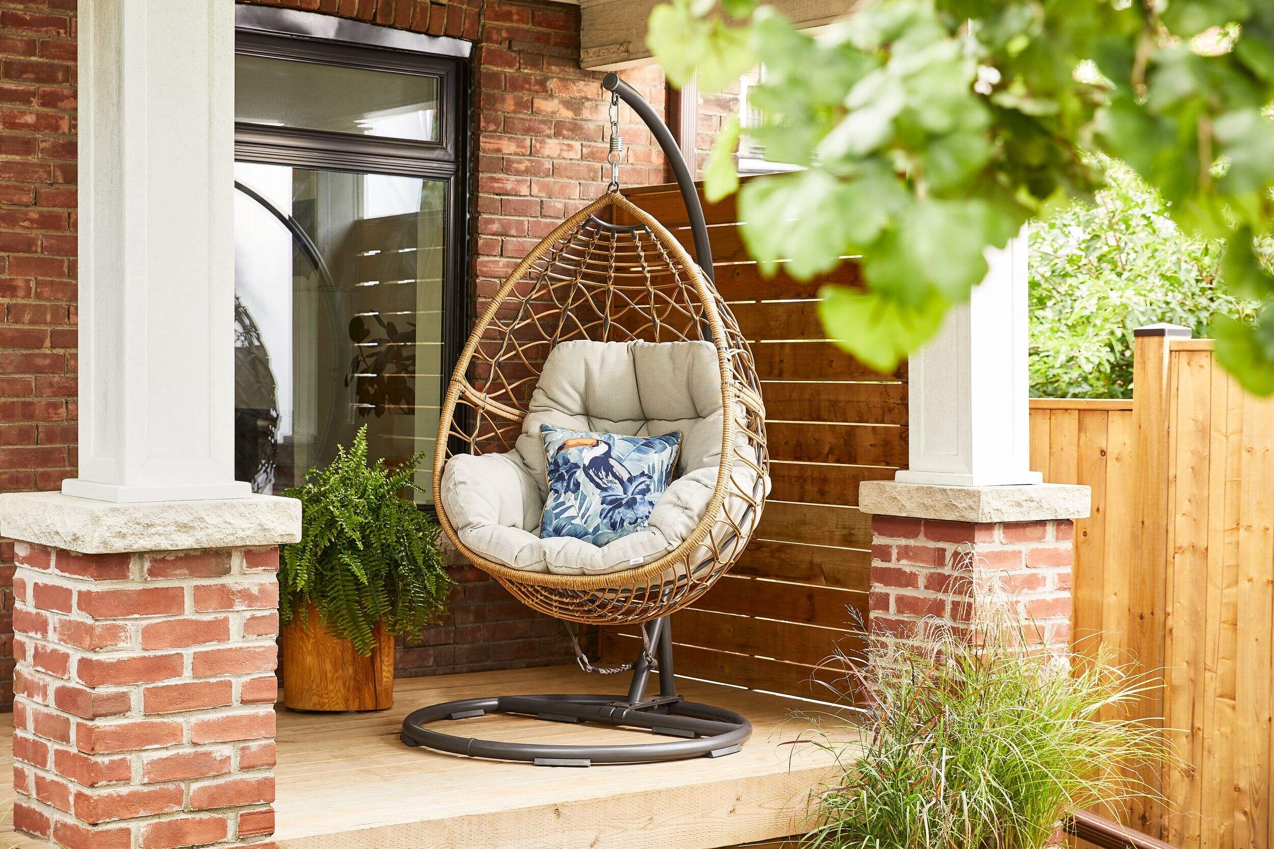 Hanging egg chair with cushions on a porch beside potted plants, set against a brick wall