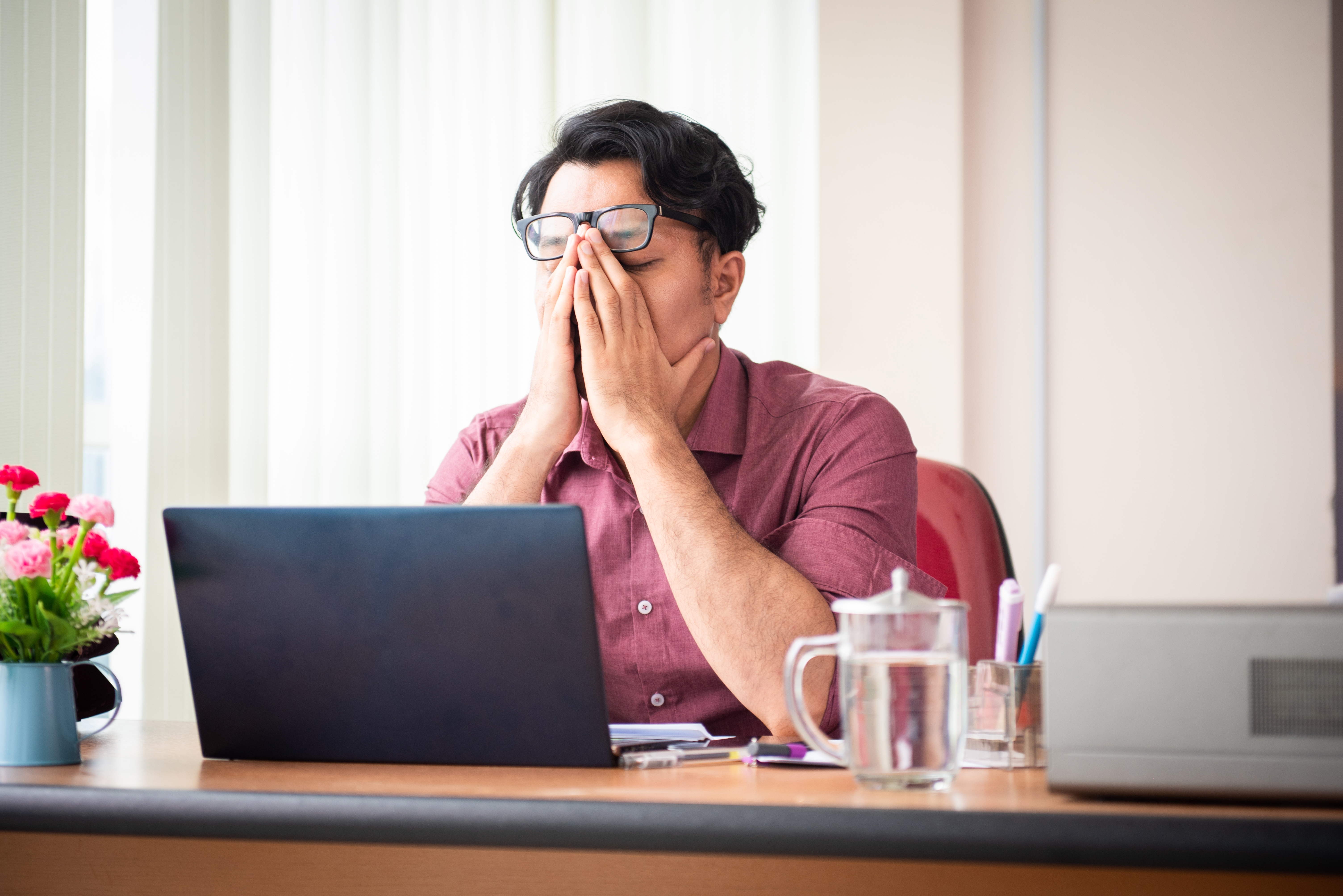 Person at a desk, rubbing their eyes in fatigue, sitting in front of a laptop and next to office supplies and a glass of water