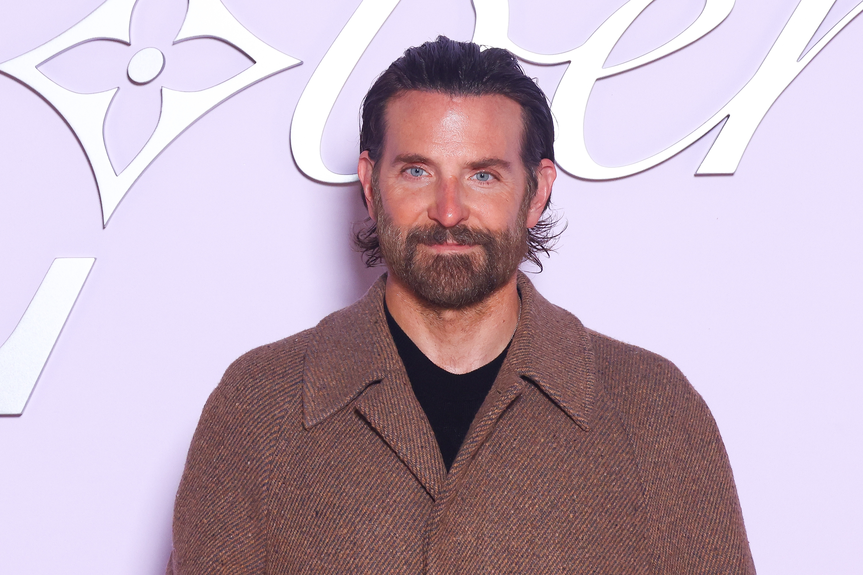 Man with a beard and long hair wearing a brown coat stands against a patterned backdrop at a celebrity event