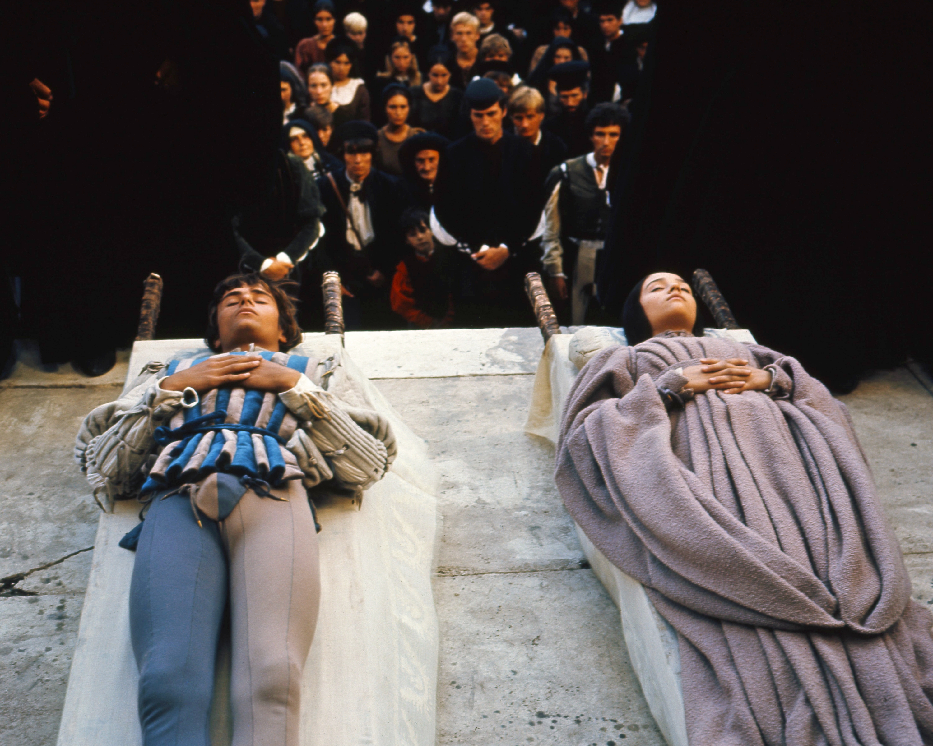 Two individuals lie side by side in a dramatic scene, surrounded by an audience in mourning attire
