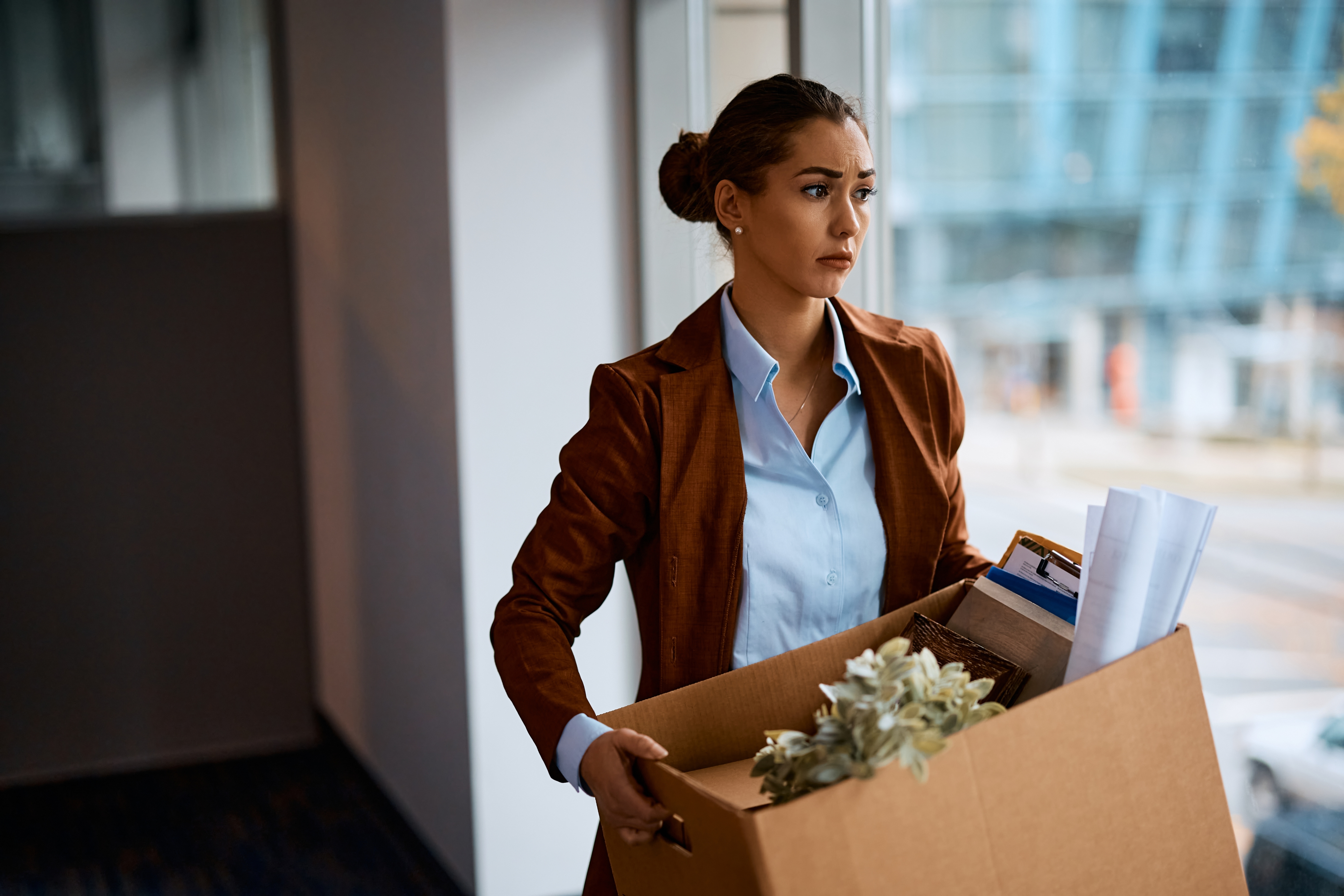 Person holding a box of office supplies, looking contemplative while standing near a window
