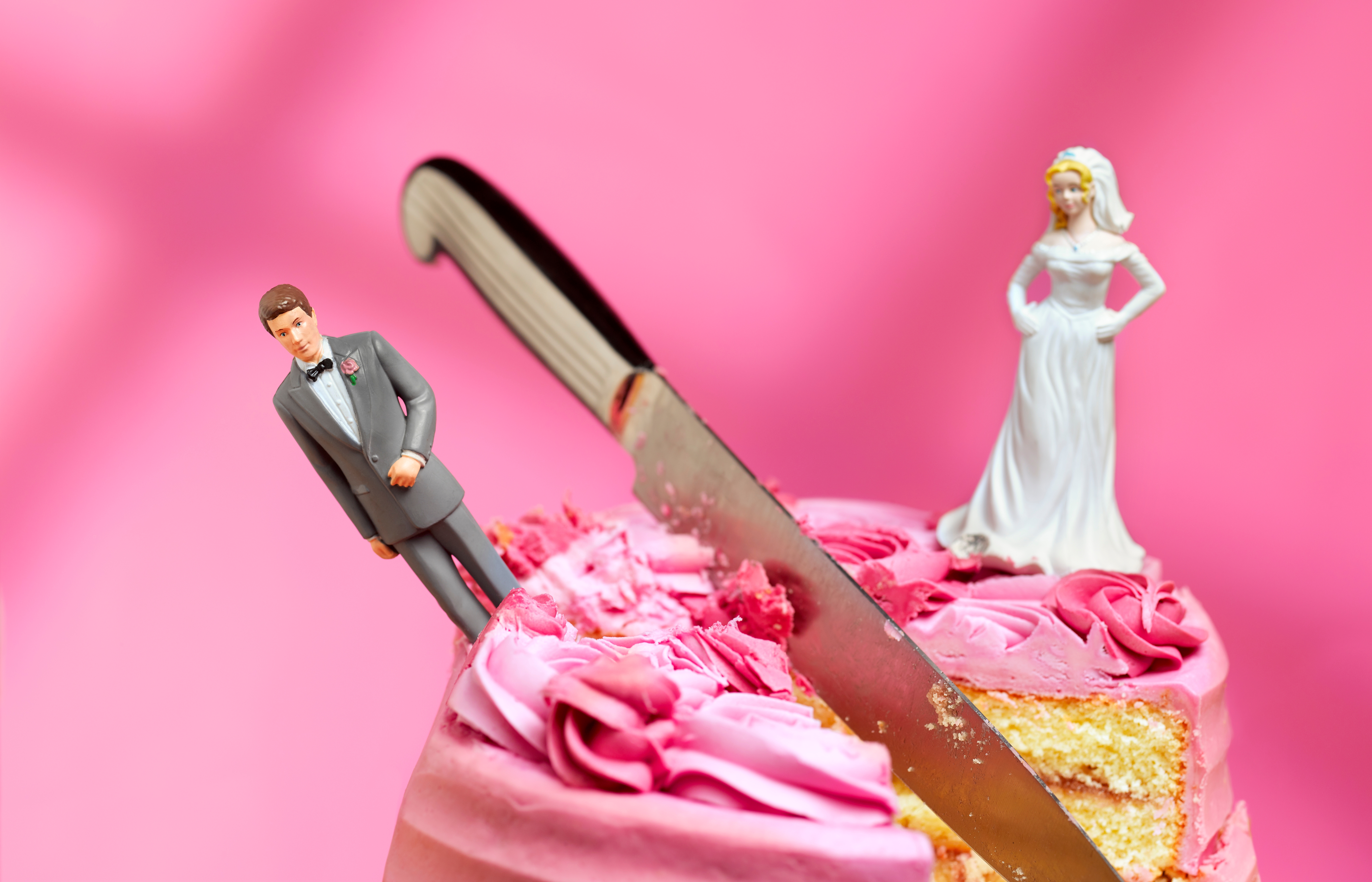 A wedding cake is topped with a plastic bride and groom. A knife is inserted into the cake between them