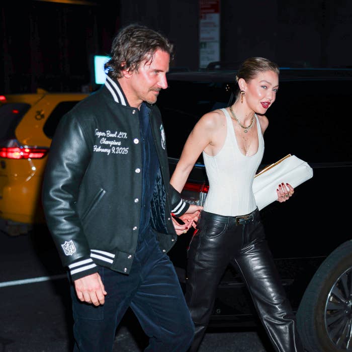 A man in a varsity jacket and a woman in a sleeveless top with leather pants are holding hands while walking past a taxi