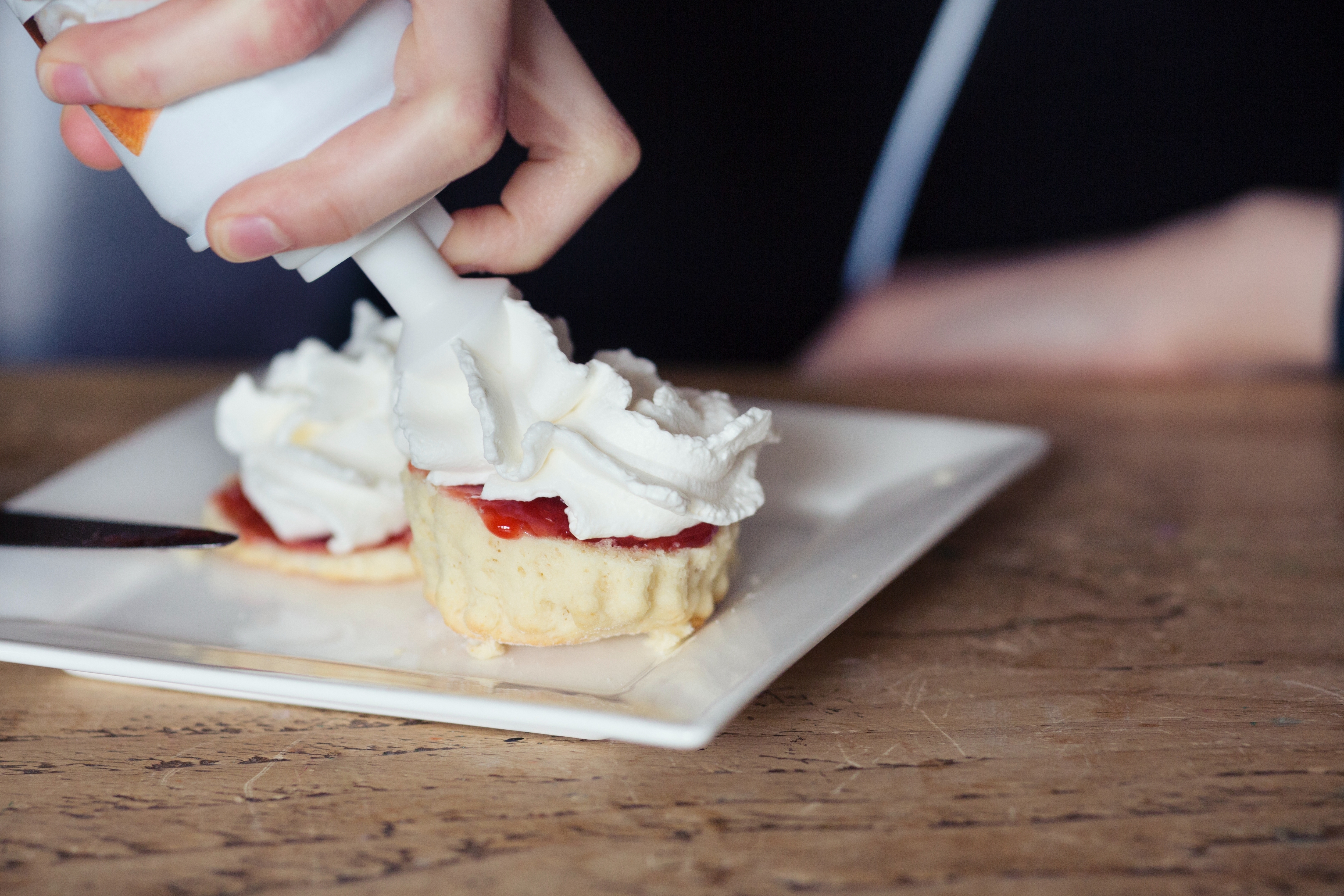 Whipped cream is being added to two biscuits with jam on a square plate