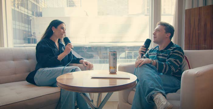 Two people seated on a sofa, holding microphones, engaged in conversation with a beverage on the coffee table between them