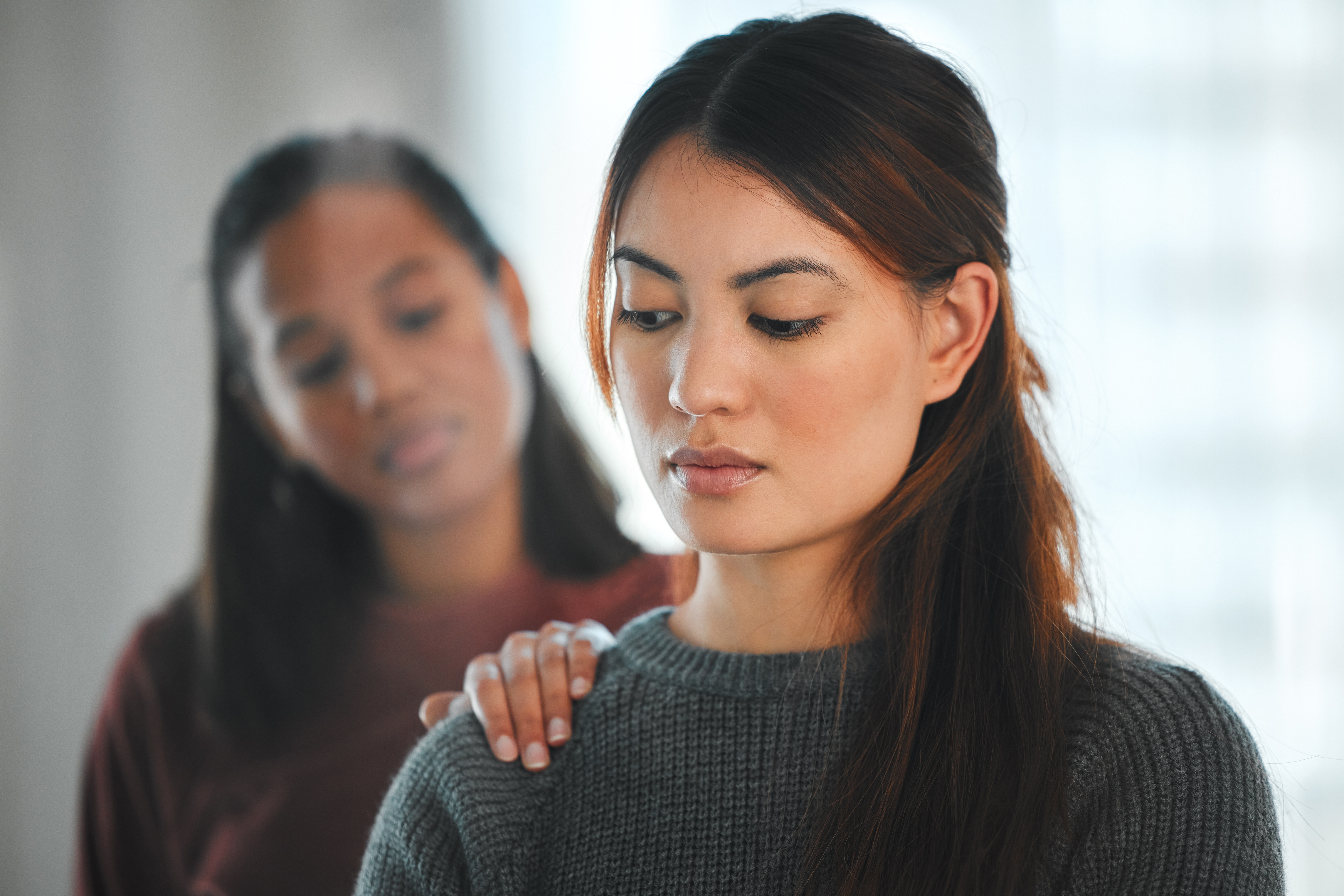 Two women are in an emotional moment. The woman in focus looks pensive, as the other woman gently rests a hand on her shoulder