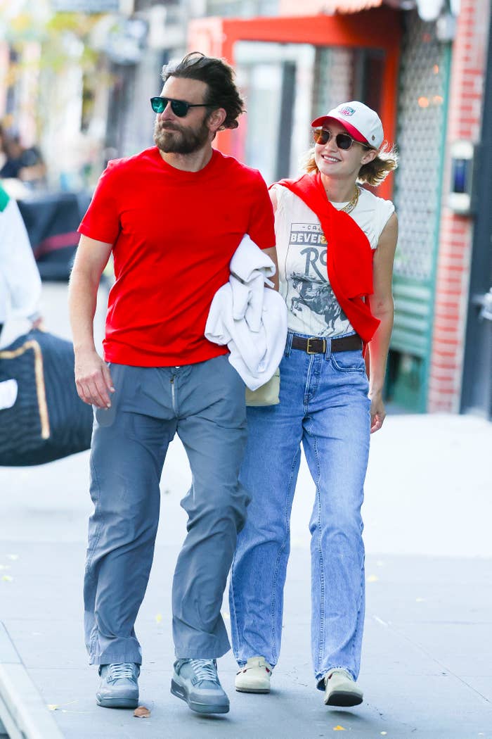Two individuals casually walk together; one wears a casual red outfit and sunglasses, the other in jeans, with a hat and sunglasses, arm linked
