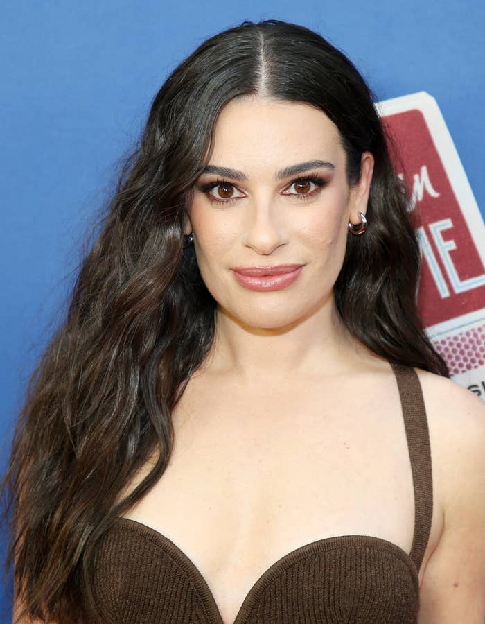 Person with long hair in a stylish top, posed against a simple blue backdrop, wearing hoop earrings