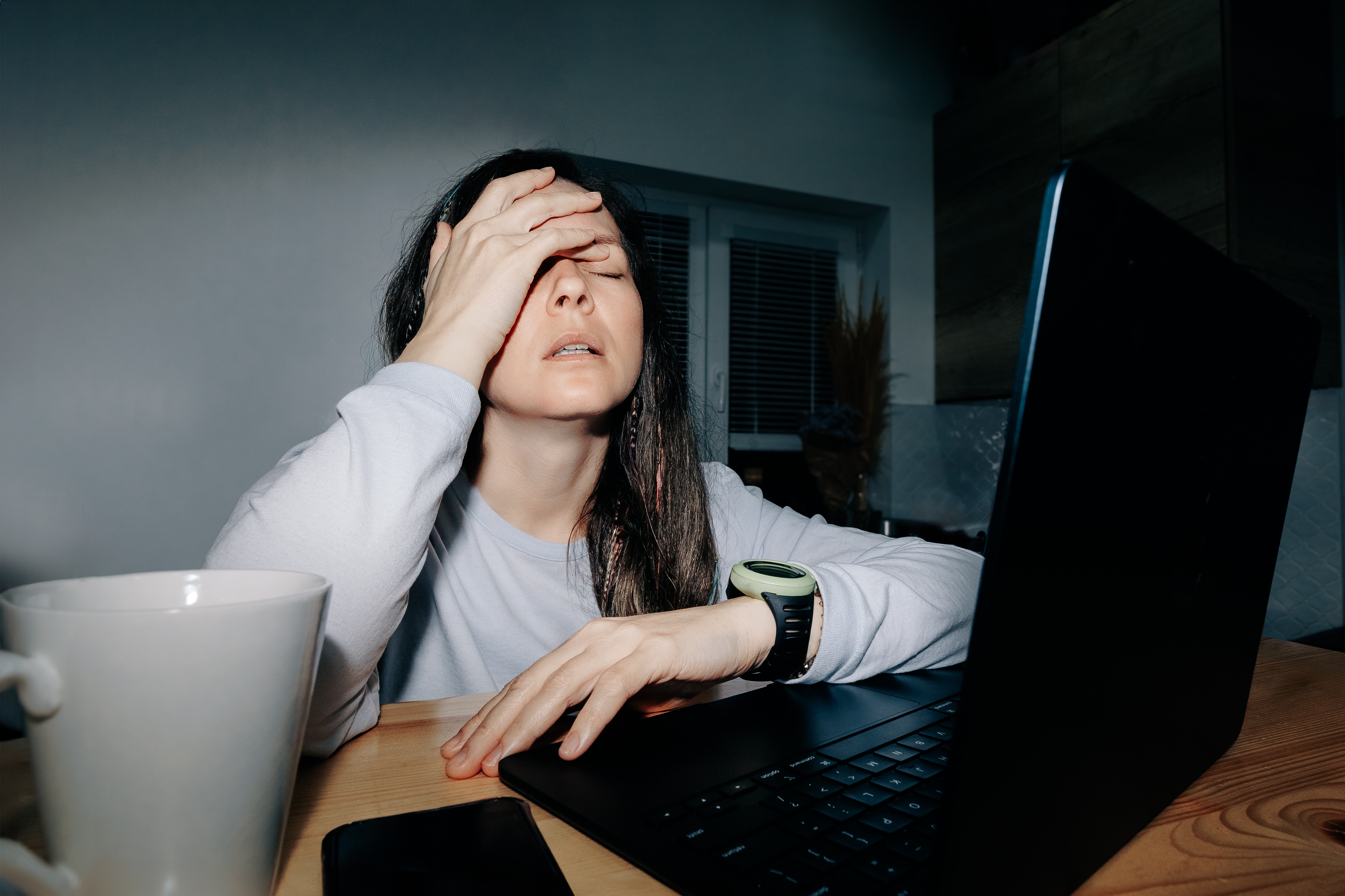 Person with hand on forehead sits at a desk with a laptop, looking stressed. A mug and smartphone are nearby