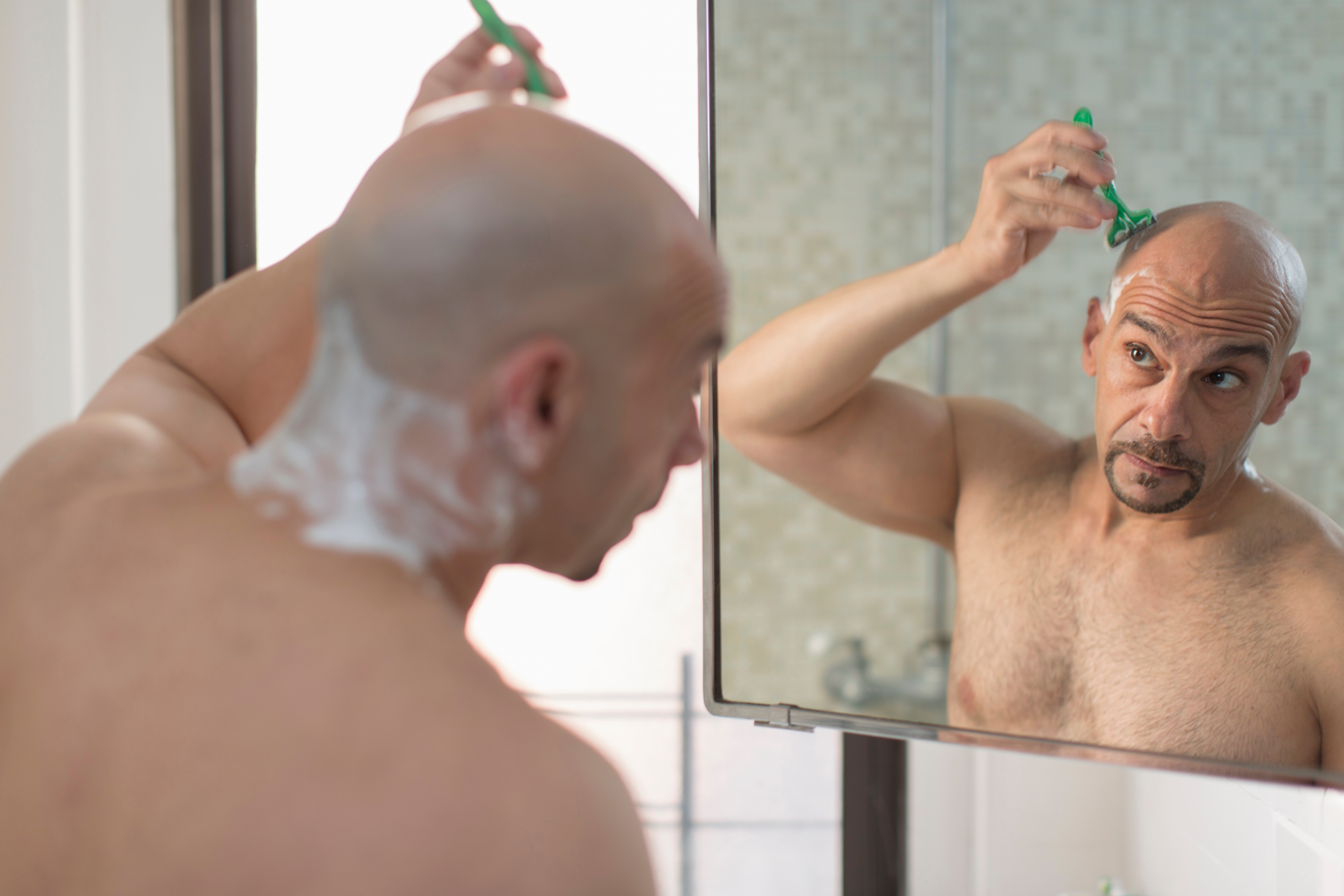 Man shaving his head with a razor, looking in a mirror; lather on his neck