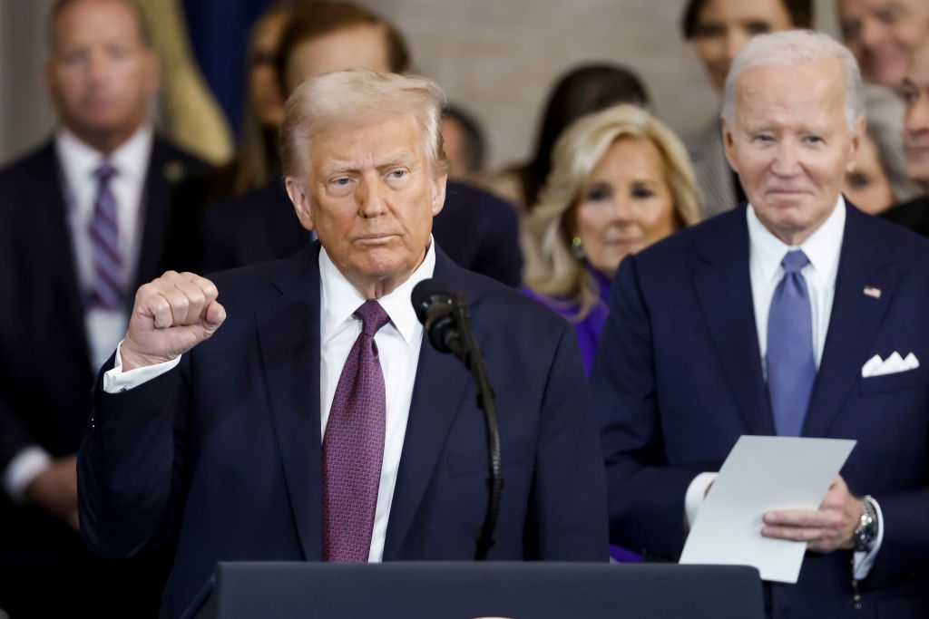 Two public figures stand on stage; one raises a fist. The other holds documents and smiles