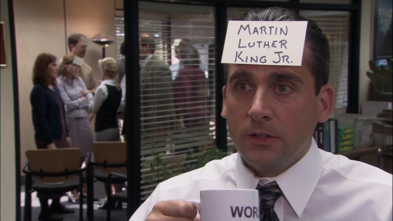 Man in an office setting with a paper reading "Martin Luther King Jr." on his forehead, holding a mug. Others are in the background