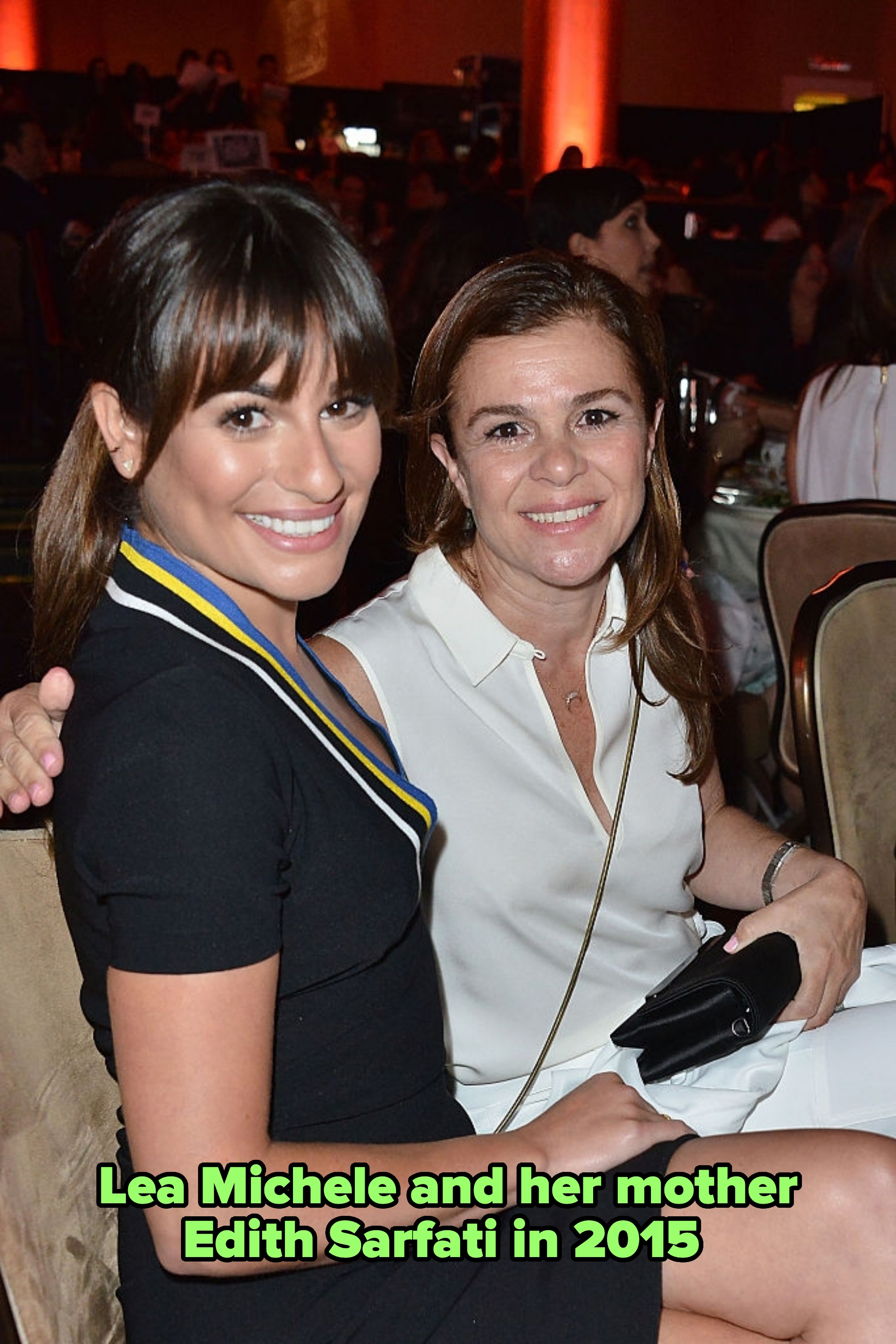 Two people smiling at a formal event. The person on the left is wearing a black dress with a colorful ribbon detail